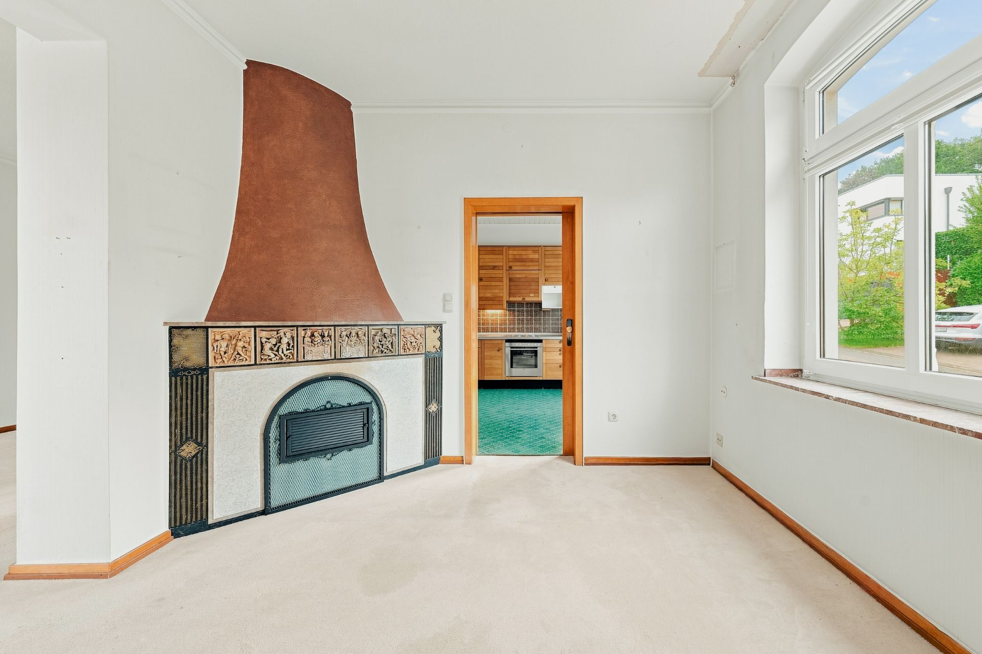 Helles Zimmer mit großem Fenster, altem Kamin und Sicht auf die angrenzende Küche mit Holzelementen.