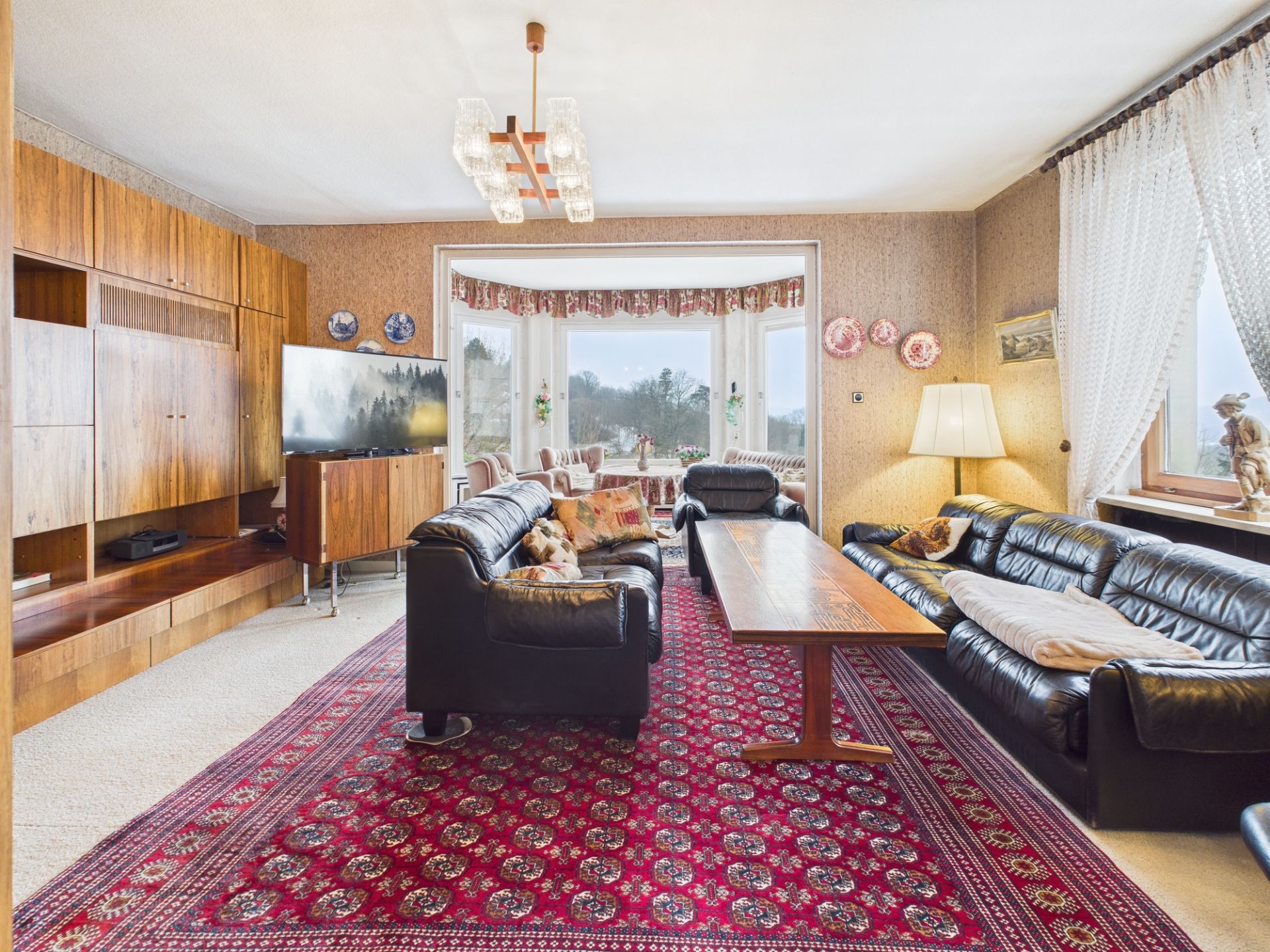 Wohnzimmer mit schwarzem Ledersofa, rotem Teppich, Holzschrankwand und großem Fenster.