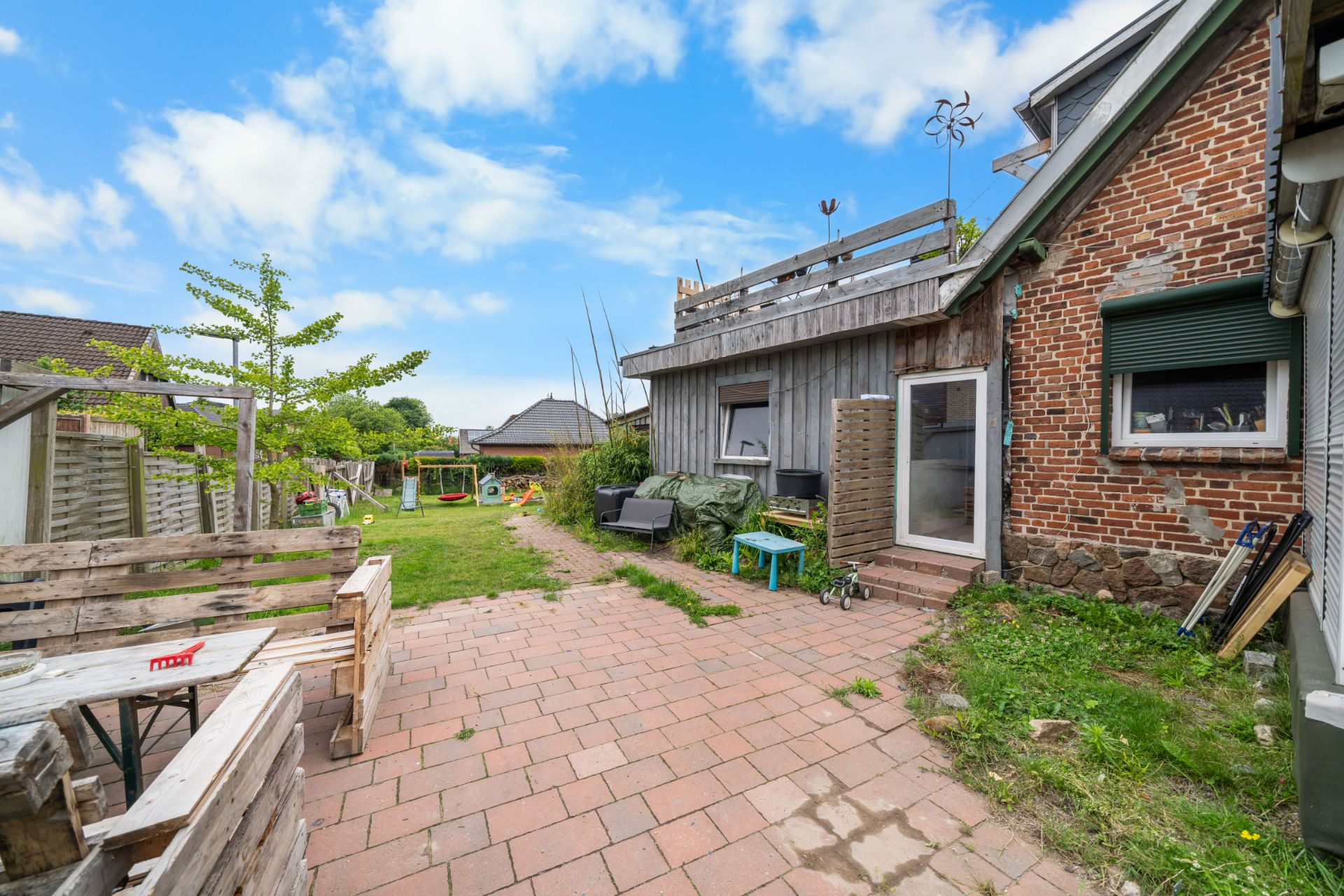 Gepflasterter Hof mit Sitzbank, Gartenmöbeln und Grünfläche neben einem Backsteinhaus.