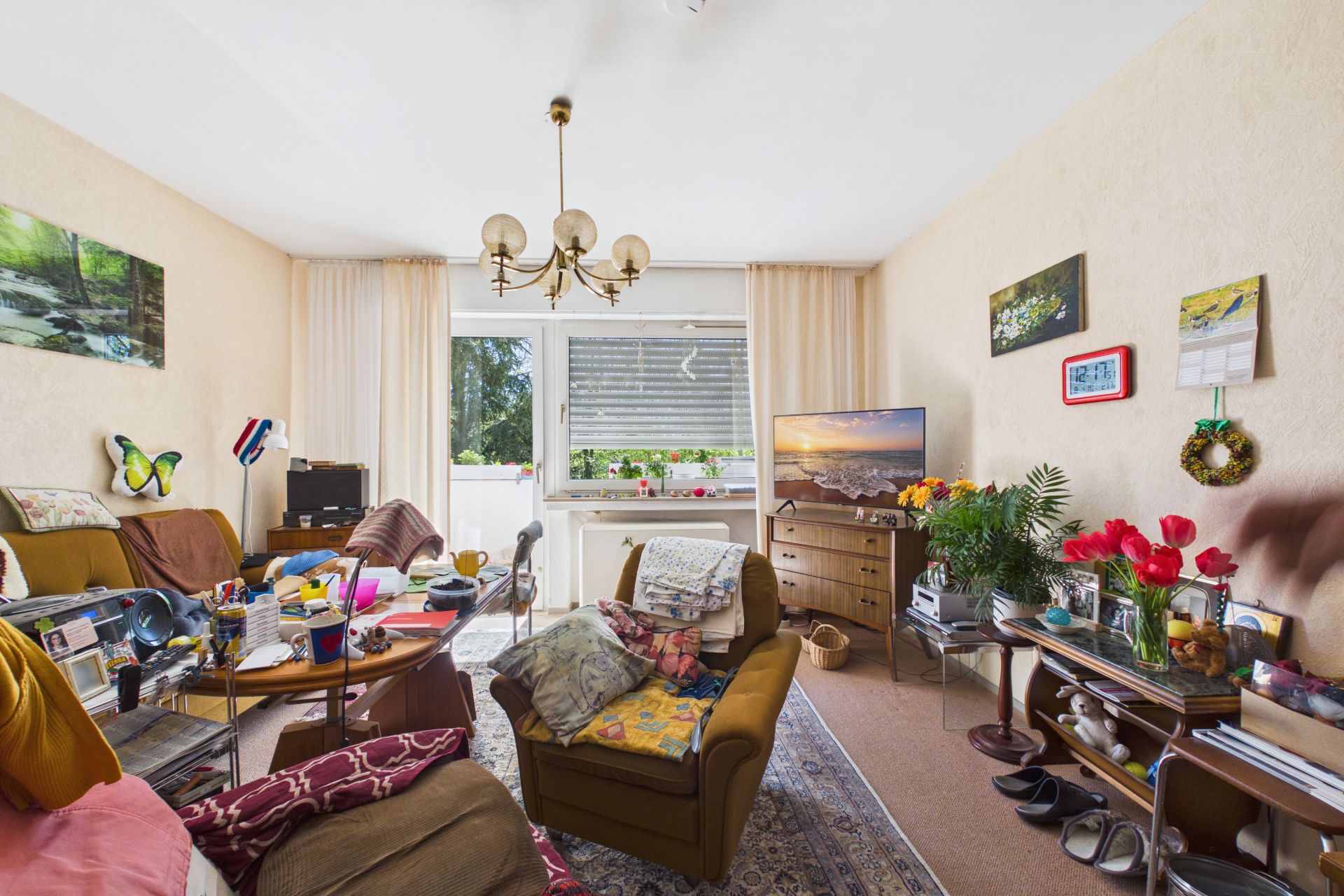 Wohnzimmer mit mehreren Sesseln, Fernseher, vielen Gegenständen und Blumen auf Tischen.