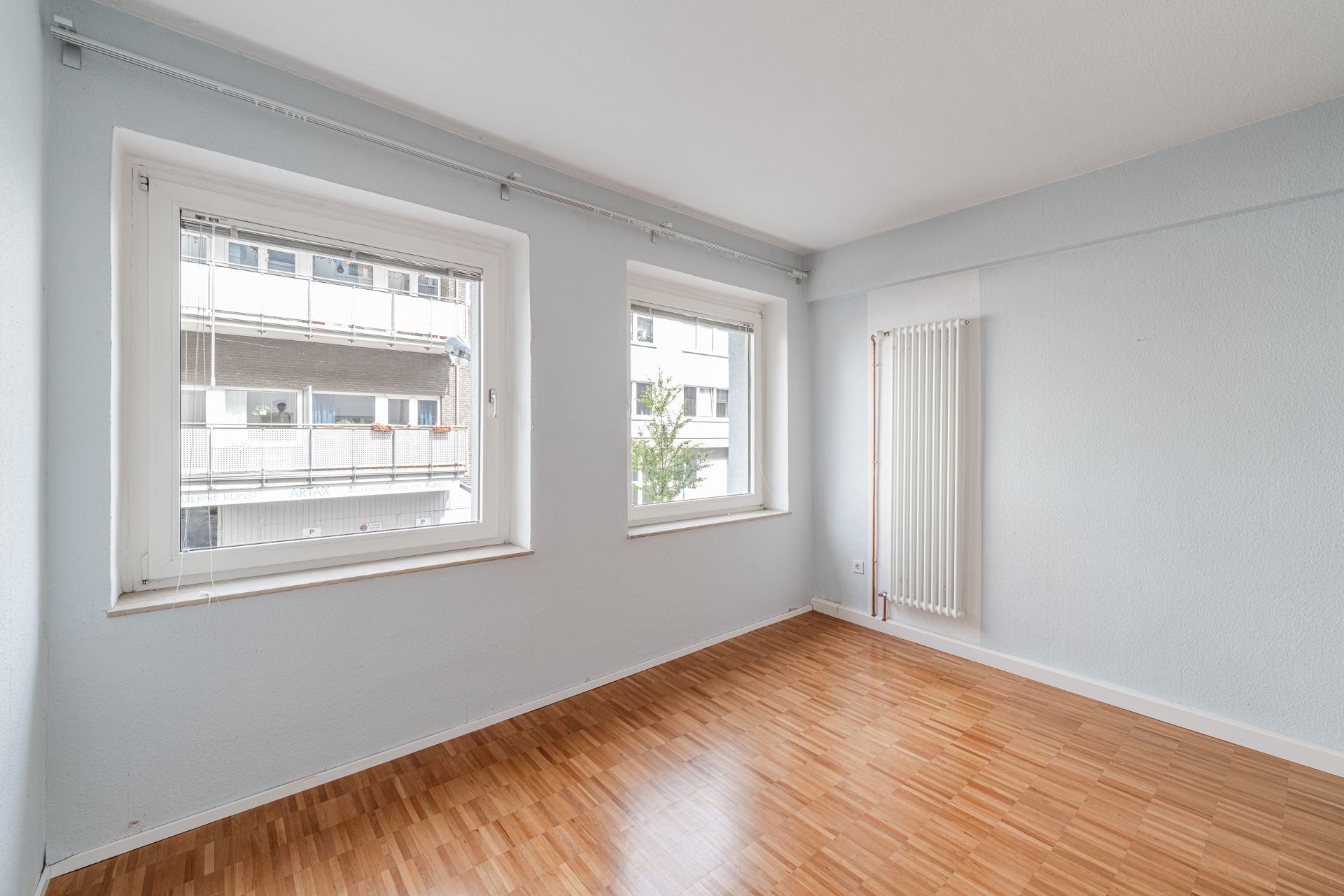 Heller, leerer Raum mit zwei Fenstern, Parkettboden und einem weißen Heizkörper an der Wand.