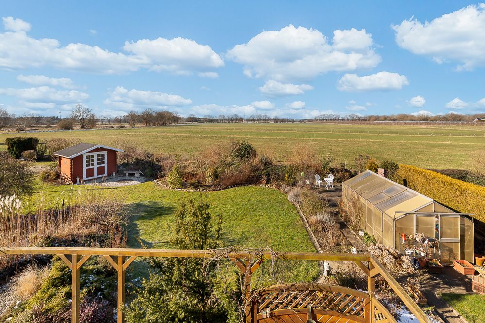 Blick auf einen großen Garten mit Gartenhaus, Gewächshaus und Sitzbereich, dahinter weite Felder.