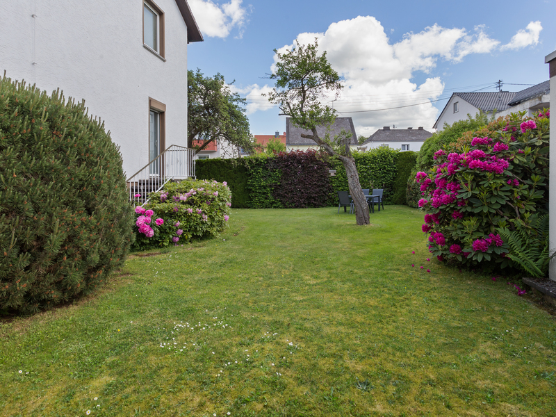 Garten mit grünem Rasen, blühenden Büschen, einem Baum und Gartenmöbeln neben einem Wohnhaus.