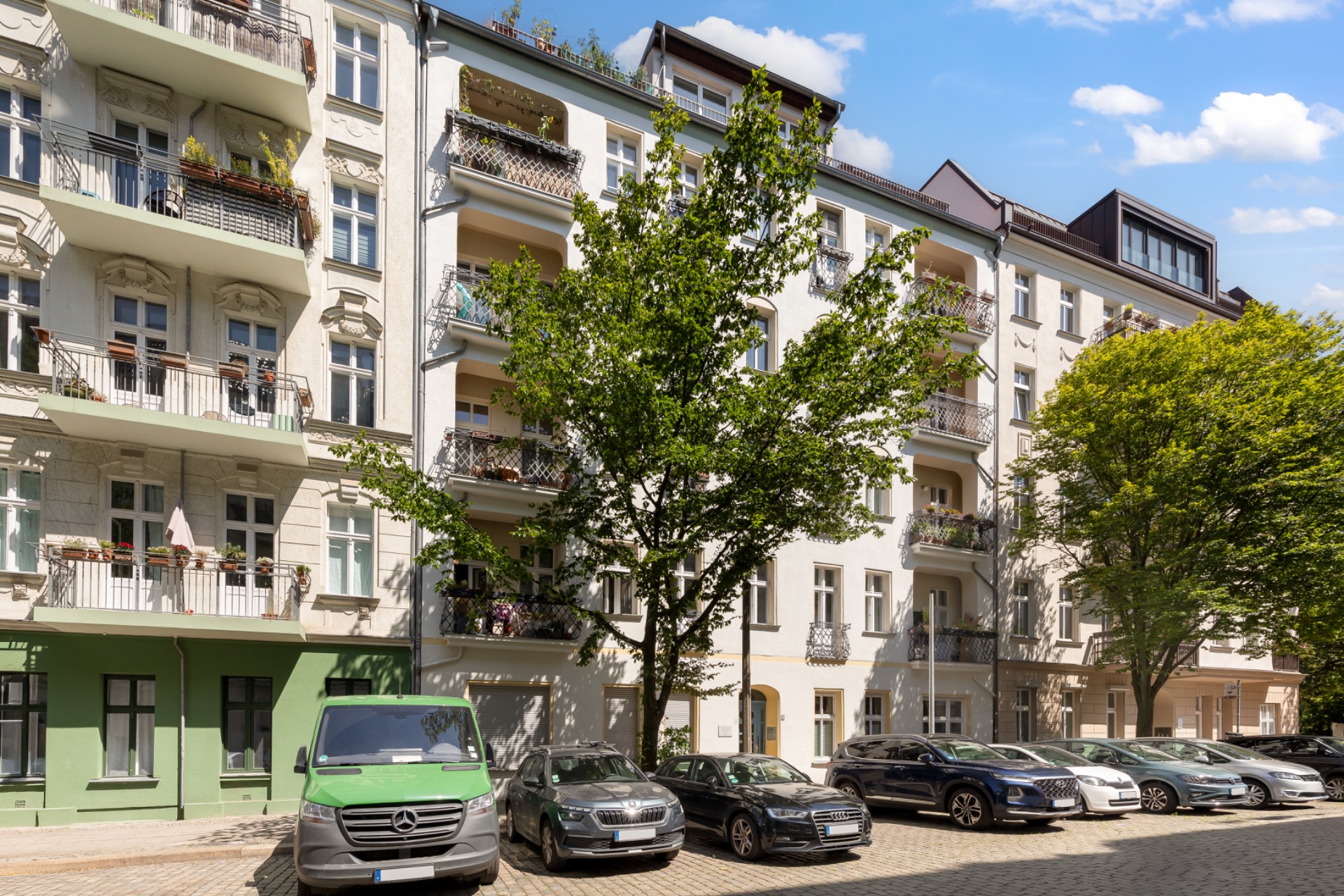 Mehrstöckiges Wohnhaus mit Balkonen, davor geparkte Autos und Bäume auf dem Gehweg.