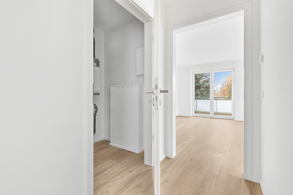 Heller Flur mit Holzboden, offener Tür zu einem Zimmer mit Balkon und großen Fenstern.