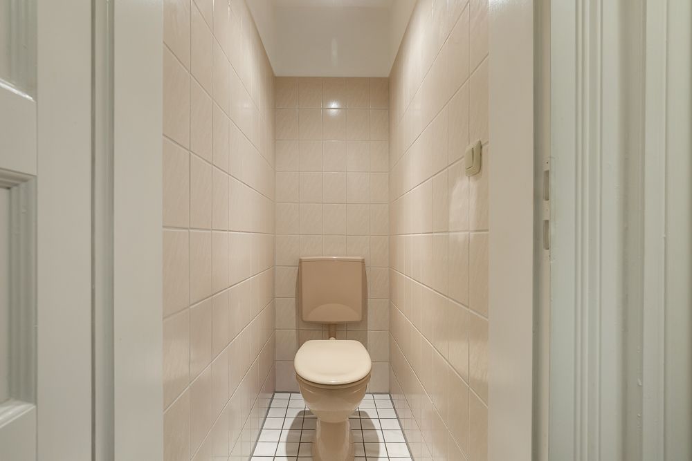 Schmaler Raum mit beigefarbenen Fliesen und einer Toilette in der Mitte.