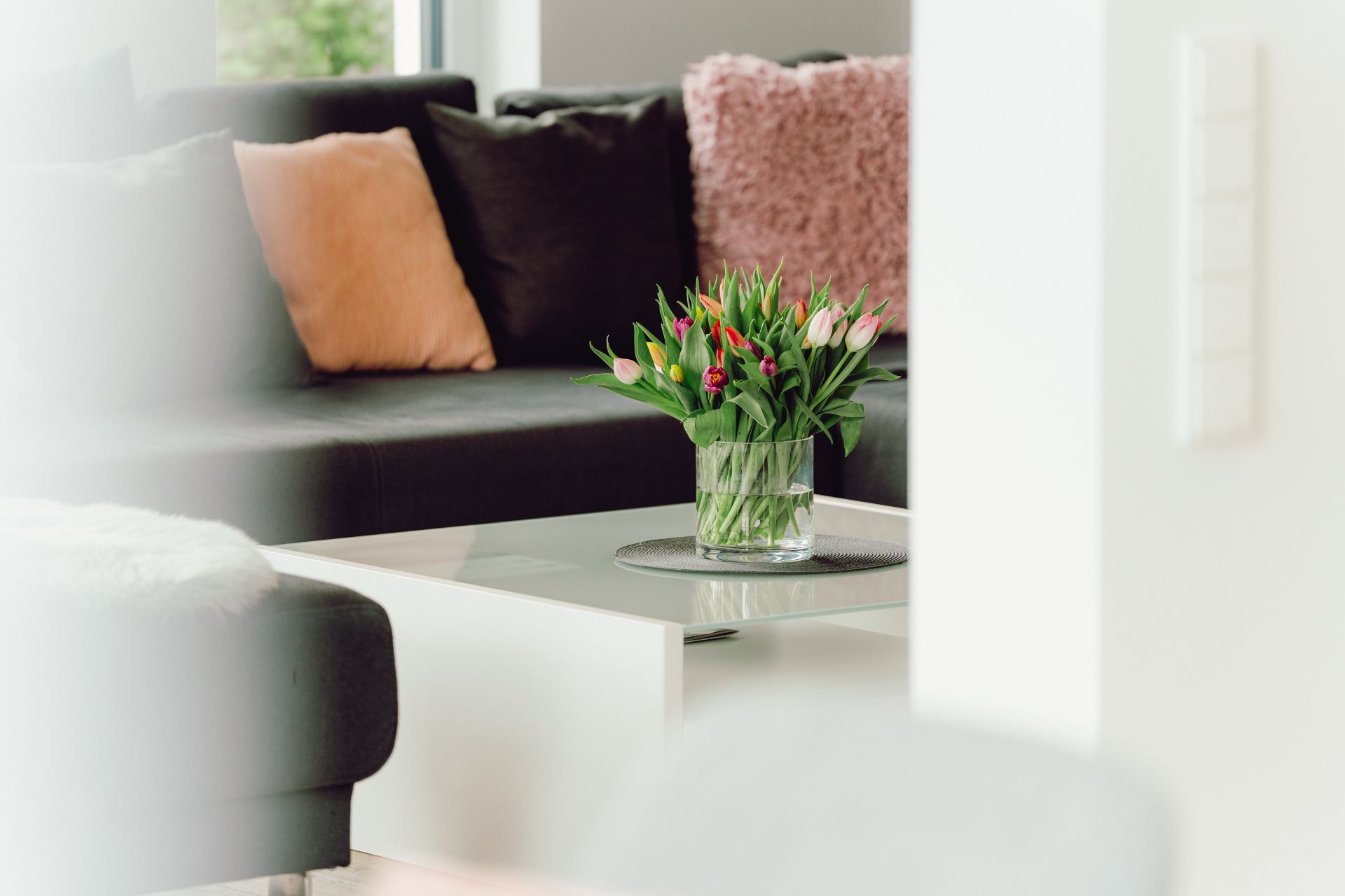 Blumenstrauß in Vase auf weißem Couchtisch vor dunklem Sofa mit Kissen.