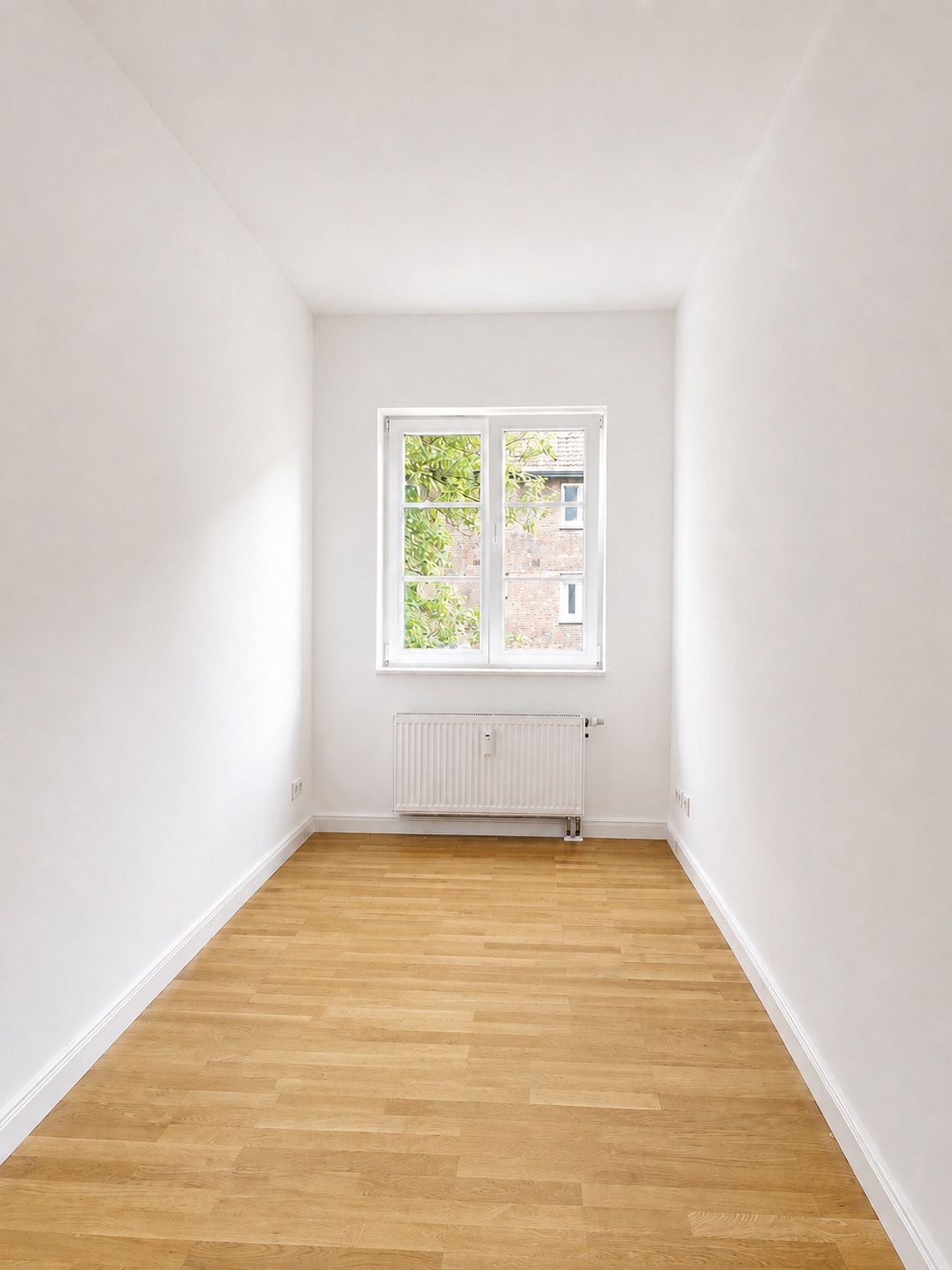 Leerer, heller Raum mit Holzfußboden und einem Fenster, darunter ein Heizkörper.