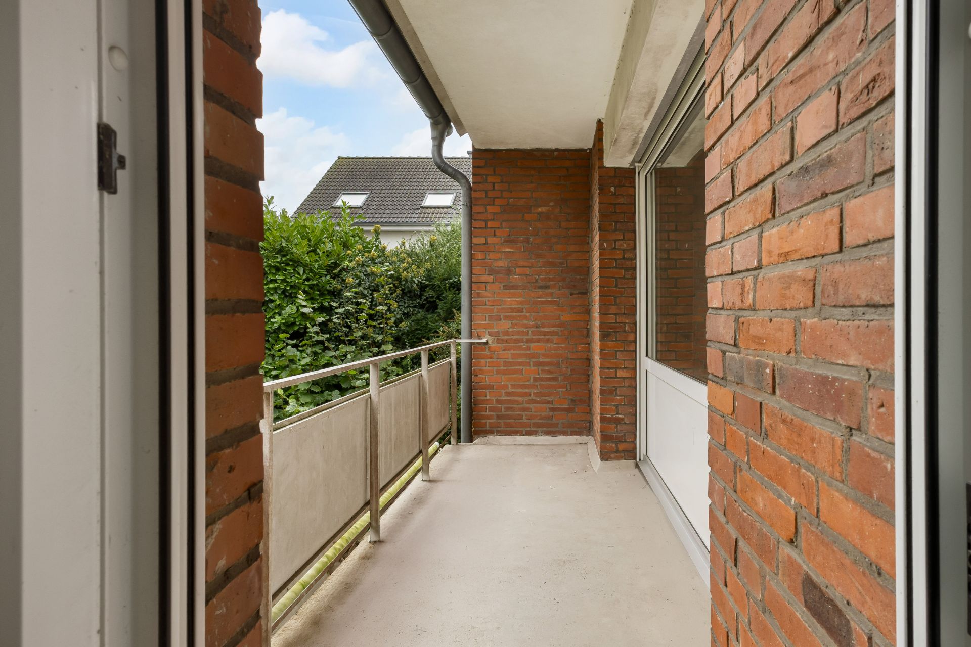 Balkon mit Geländer, Backsteinwänden und Blick auf grüne Hecke.