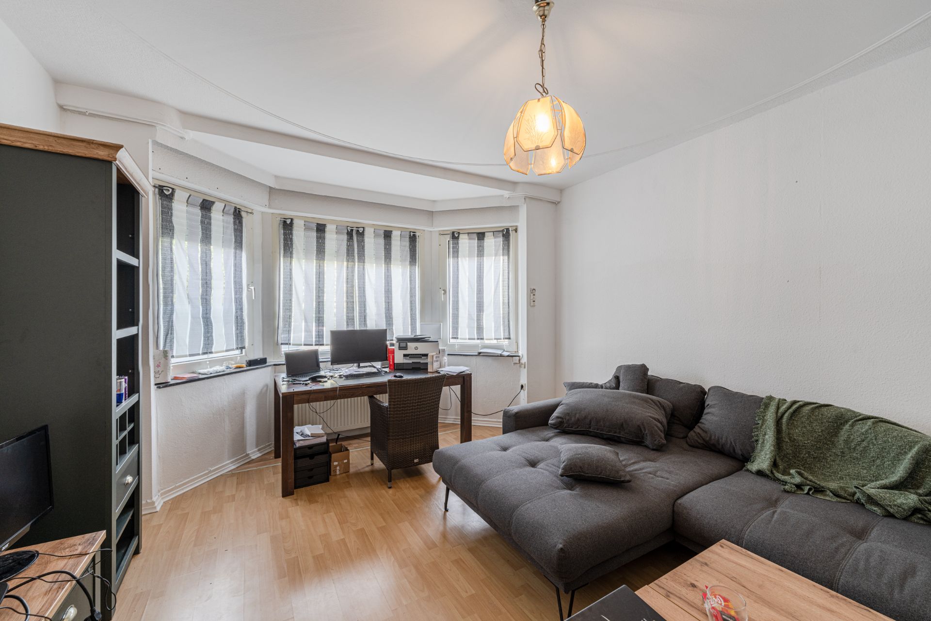 Wohnzimmer mit grauem Ecksofa, Schreibtisch am Fenster und Deckenleuchte.