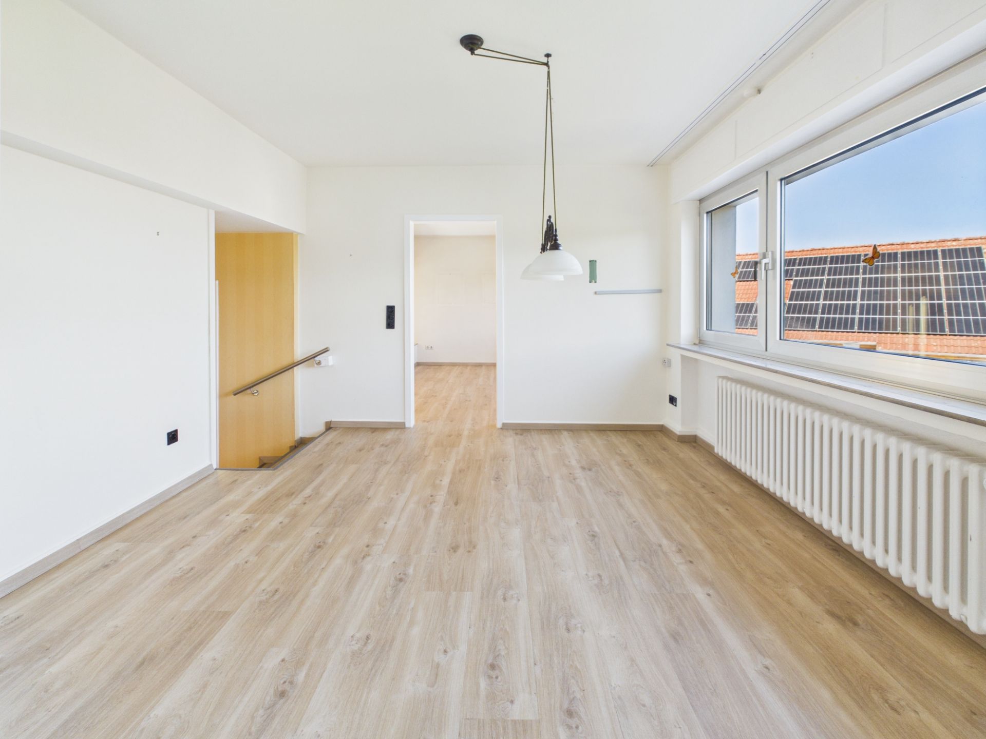 Leerer, heller Raum mit Holzfußboden, großen Fenstern und Hängelampe.