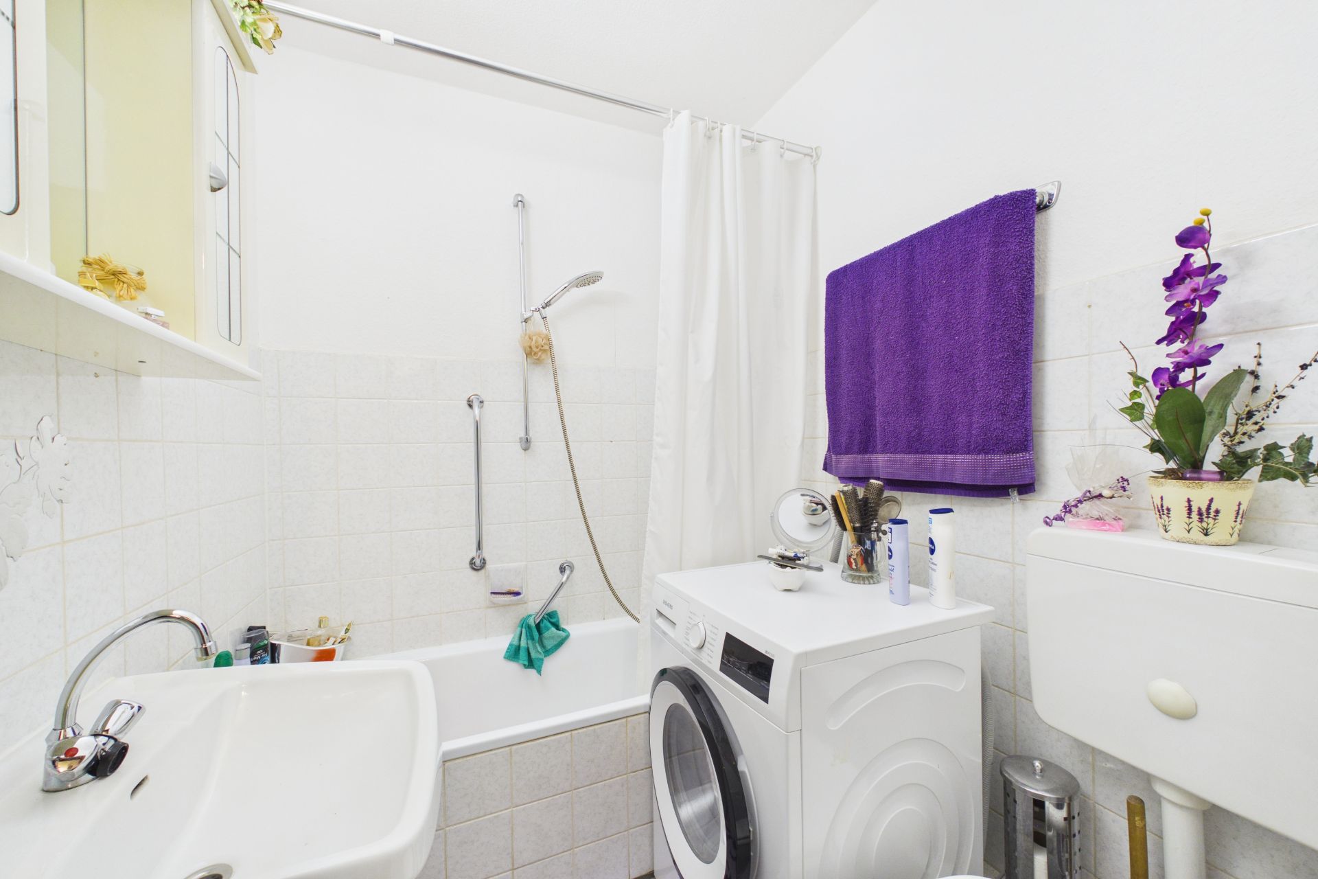 Kleines Badezimmer mit Badewanne, Waschmaschine, Waschbecken und lila Handtuch an der Wand.