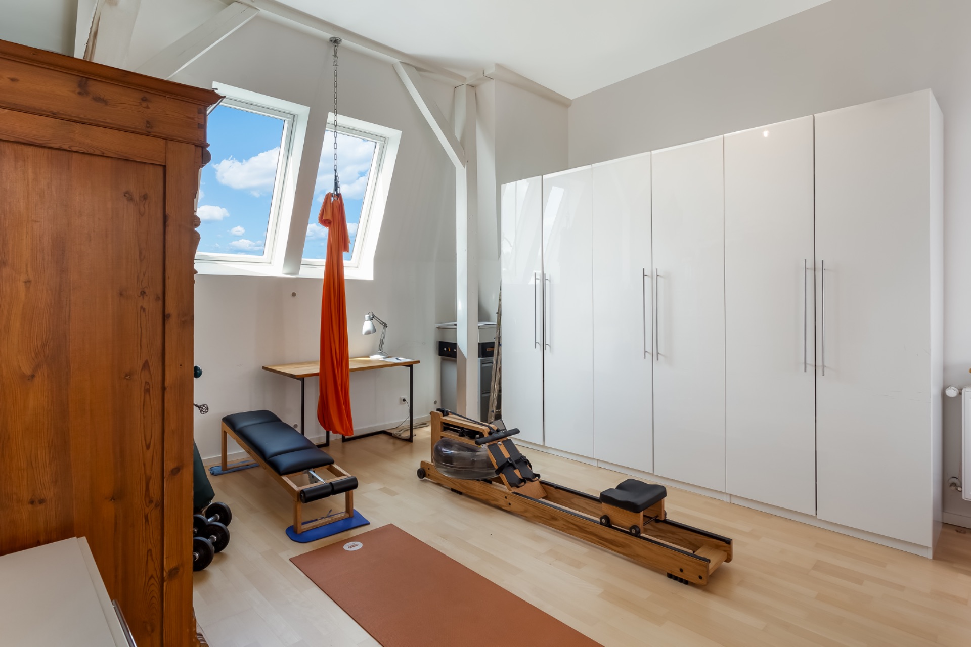 Helles Zimmer mit Sportgeräten, weißen Schränken, Holzboden und zwei Dachfenstern.