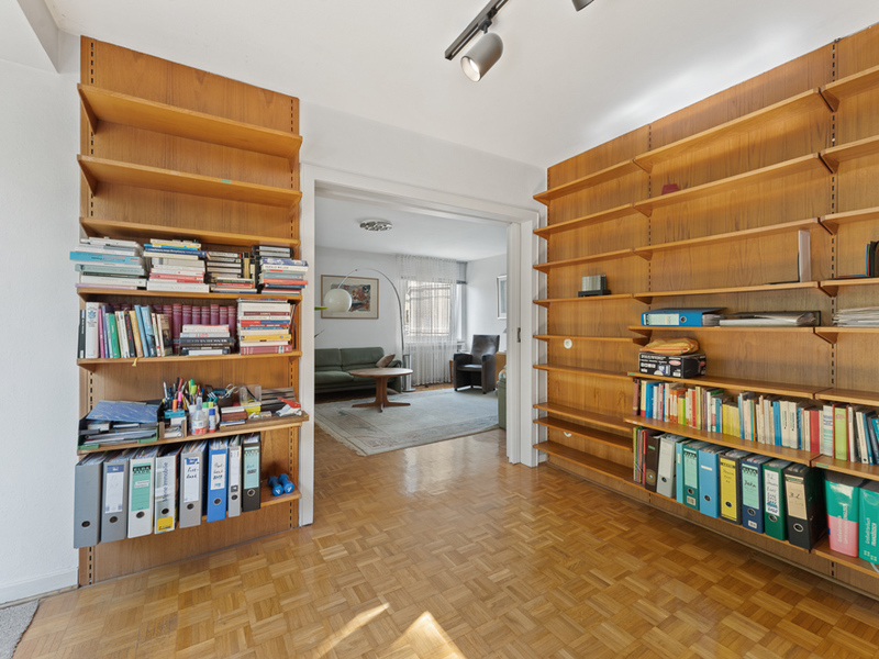 Zimmer mit Holzregalen voller Bücher und Ordner, Blick in ein weiteres Wohnzimmer mit Sofa.