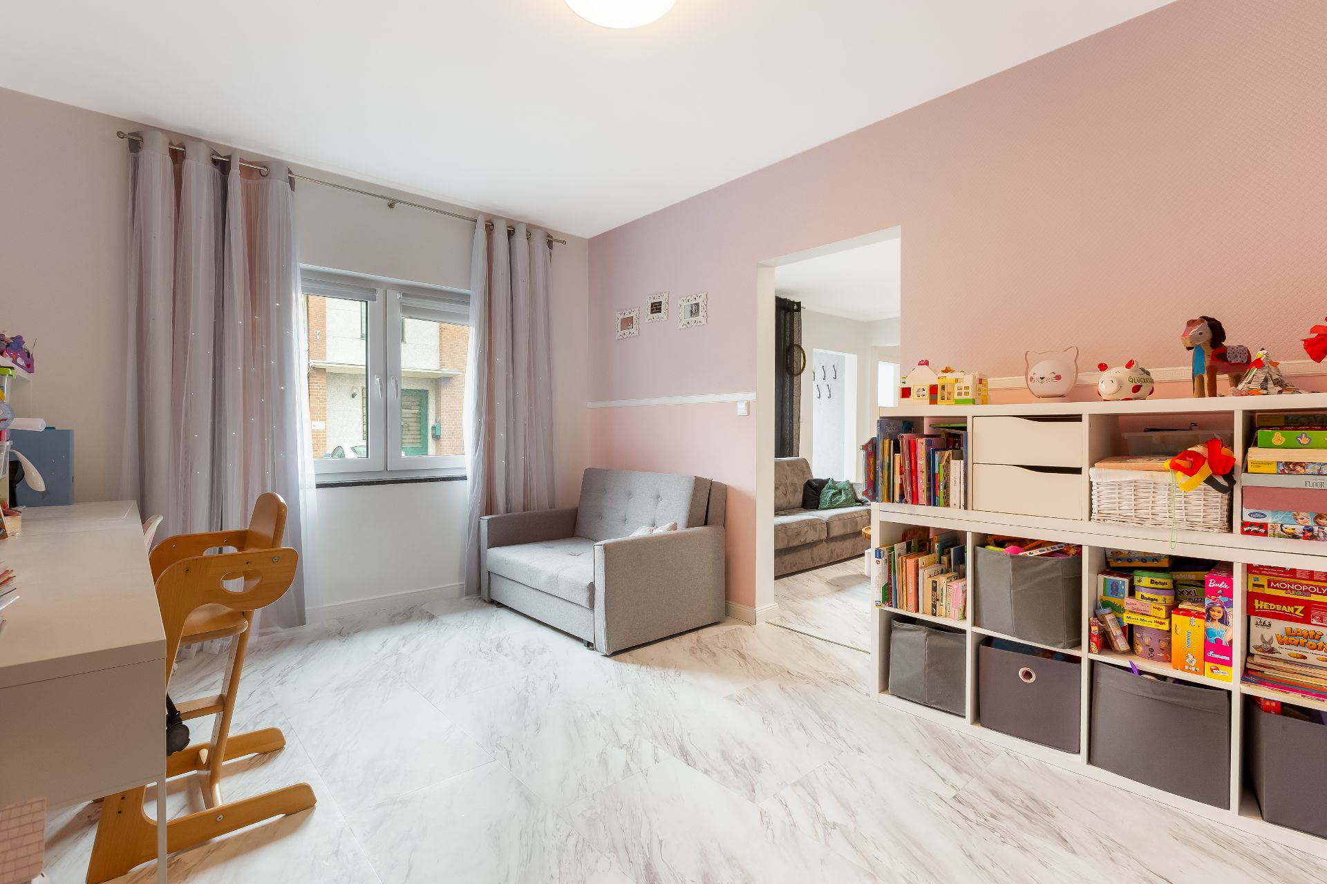Helles Kinderzimmer mit Schreibtisch, Stühlen, Sofa, Regal voller Bücher und Spielzeug.