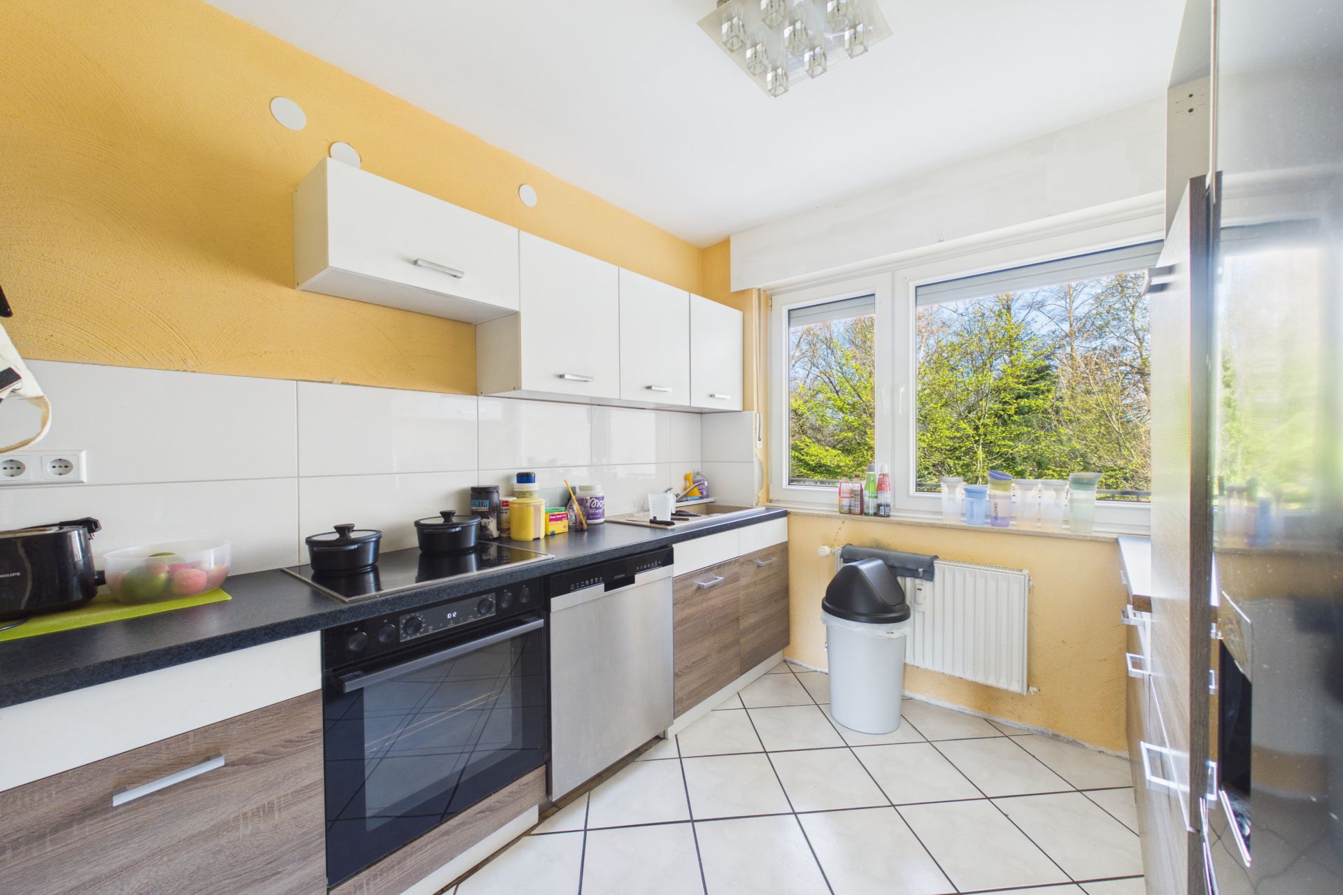 Moderne Küche mit gelben Wänden, weißen Schränken und großen Fenstern mit Blick ins Grüne.