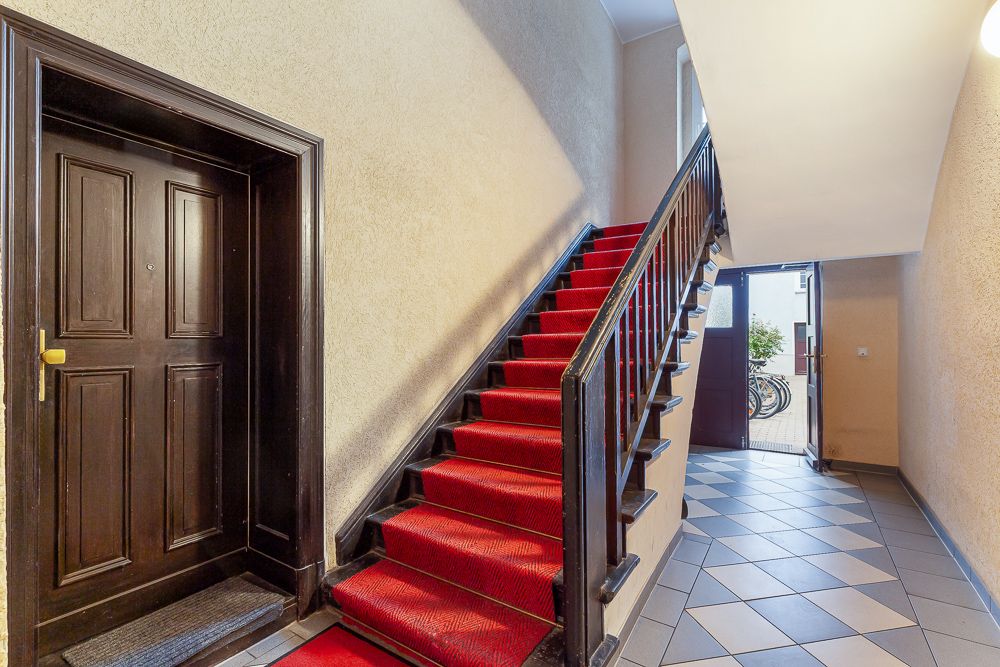 Treppenhaus mit rotem Teppich, dunkler Tür links und Fliesenboden, Eingangstür hinten offen.