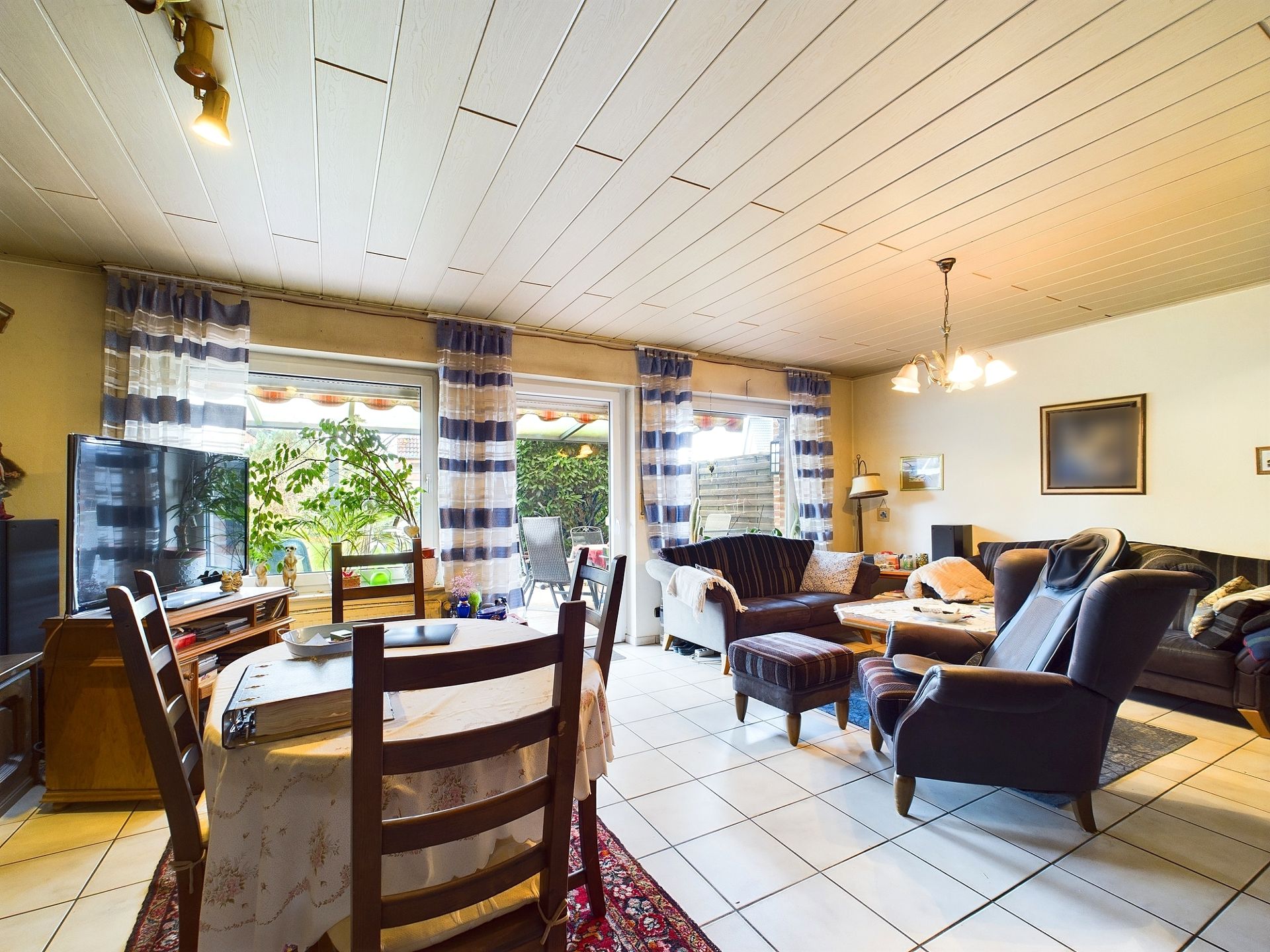 Helles Wohnzimmer mit Sofas, Sessel, Esstisch, TV und großen Fenstern zum Garten.