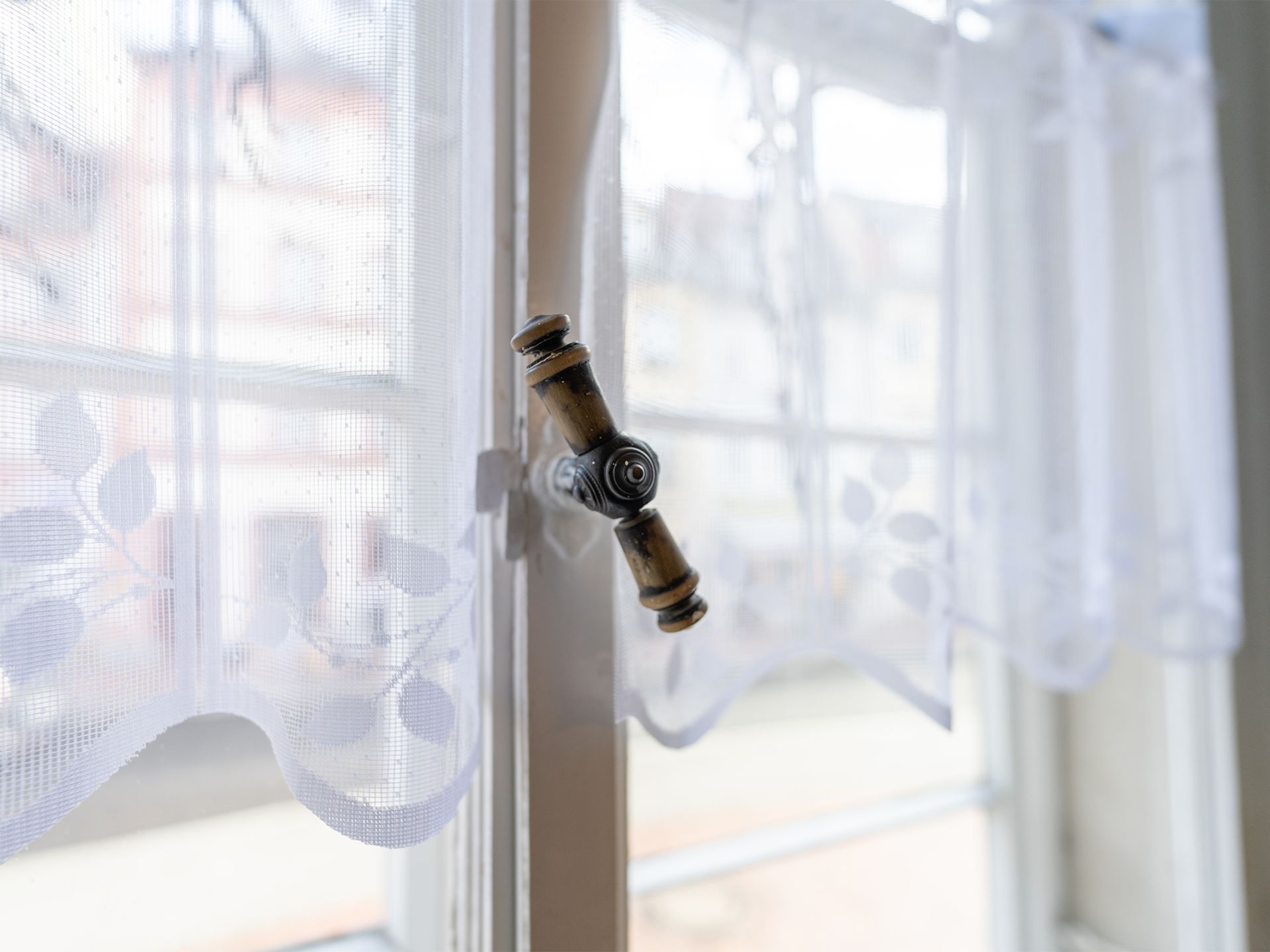 Nahaufnahme eines verzierten Fenstergriffs und weißer Gardinen mit Blumenmuster.