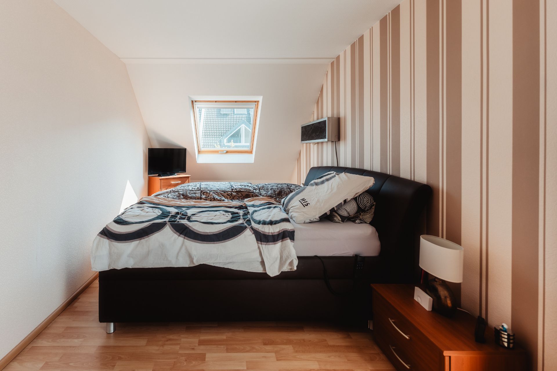 Schlafzimmer mit Doppelbett, Nachttisch, Lampe und Dachfenster, daneben ein Fernseher.
