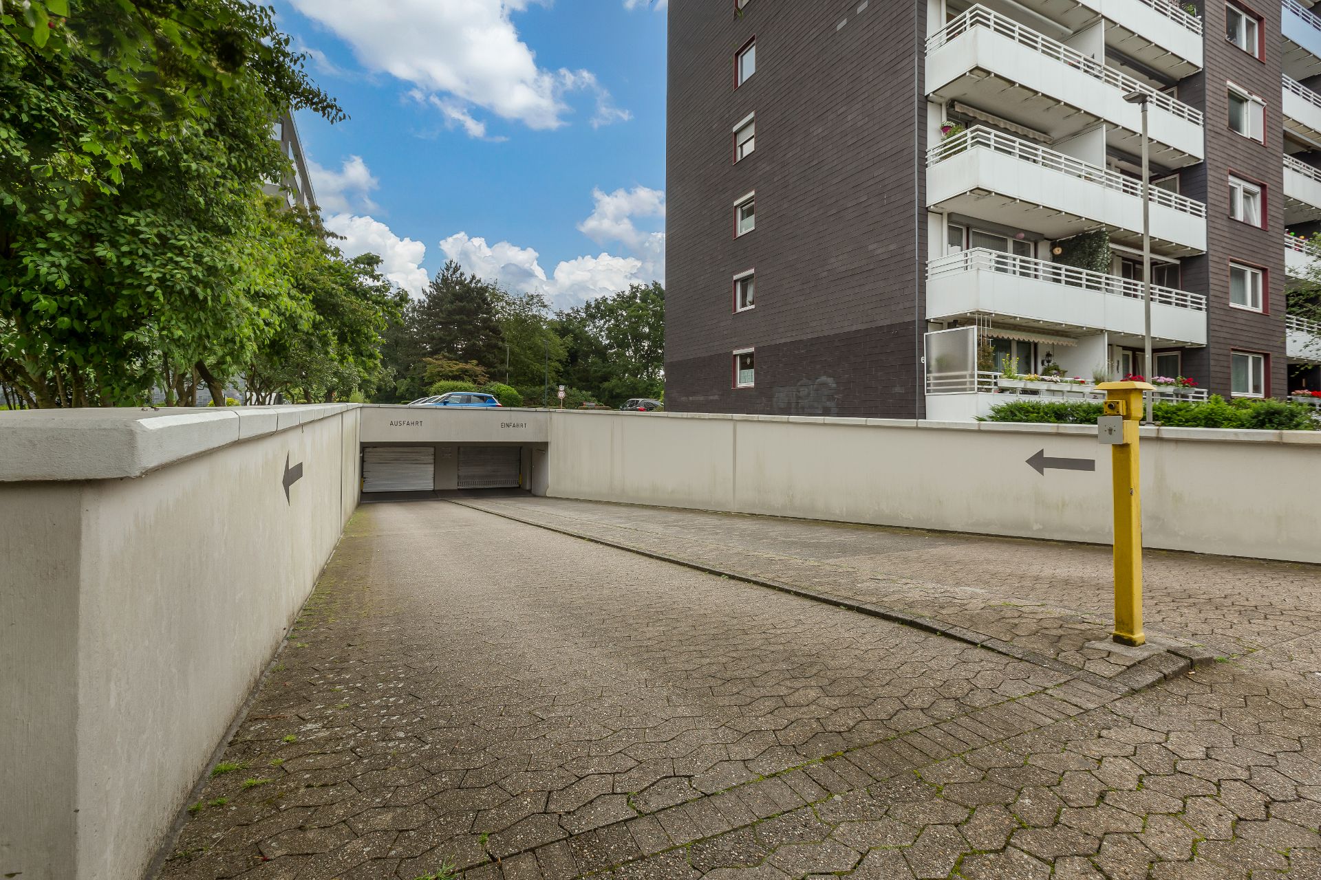 Abfahrt zur Tiefgarage neben Wohnhaus, gelbe Schranke und Bäume im Hintergrund.