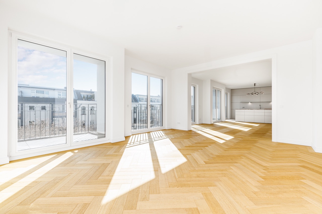 Helles Wohnzimmer mit Parkettboden, großen Fenstern und Zugang zur offenen Küche.
