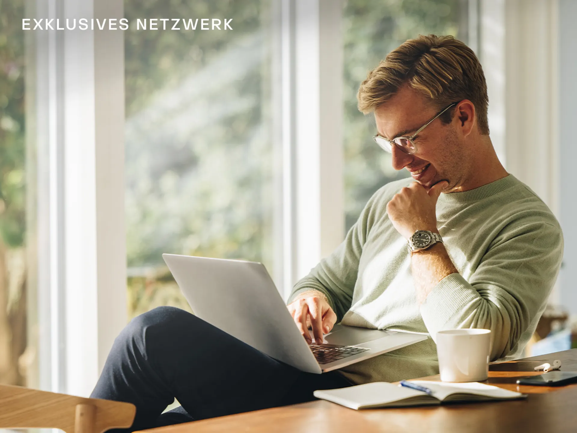 Mann sitzt am Tisch mit Laptop, Tasse, Notizbuch und Stift vor großen Fenstern.