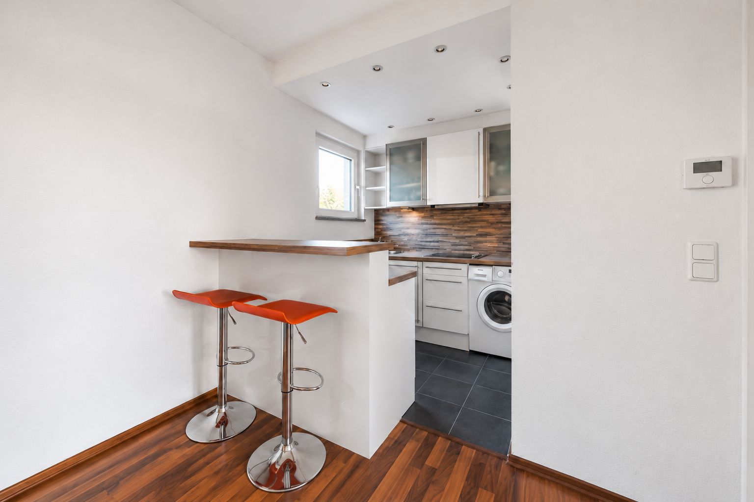 Moderne kleine Küche mit Bartresen, zwei roten Hockern, Waschmaschine und Fenster.