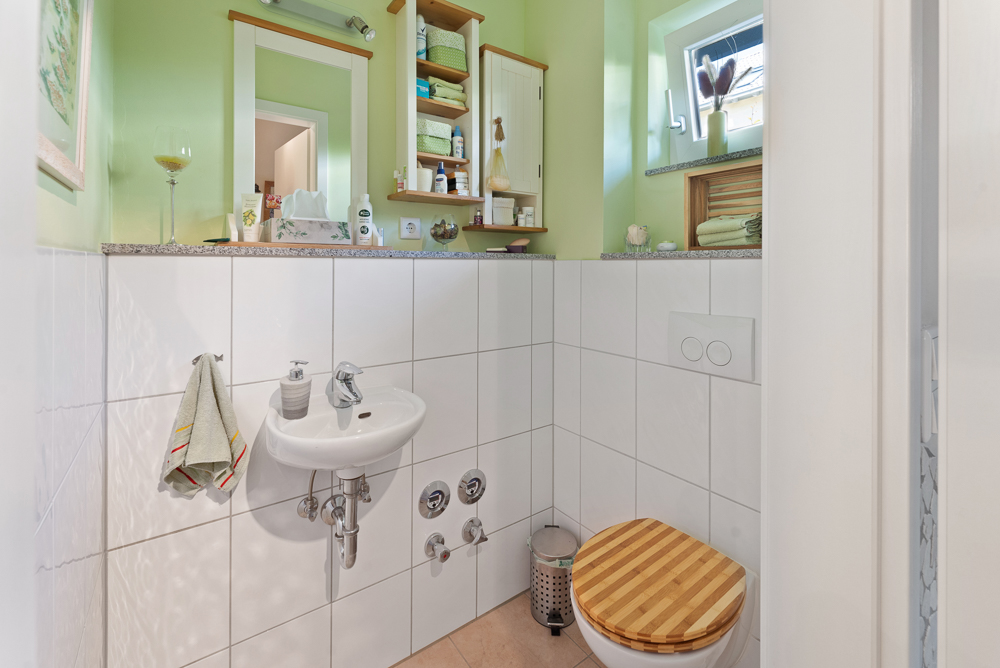 Kleines Badezimmer mit Waschbecken, Toilette und Regal, grüne Wände, weiß gefliest.