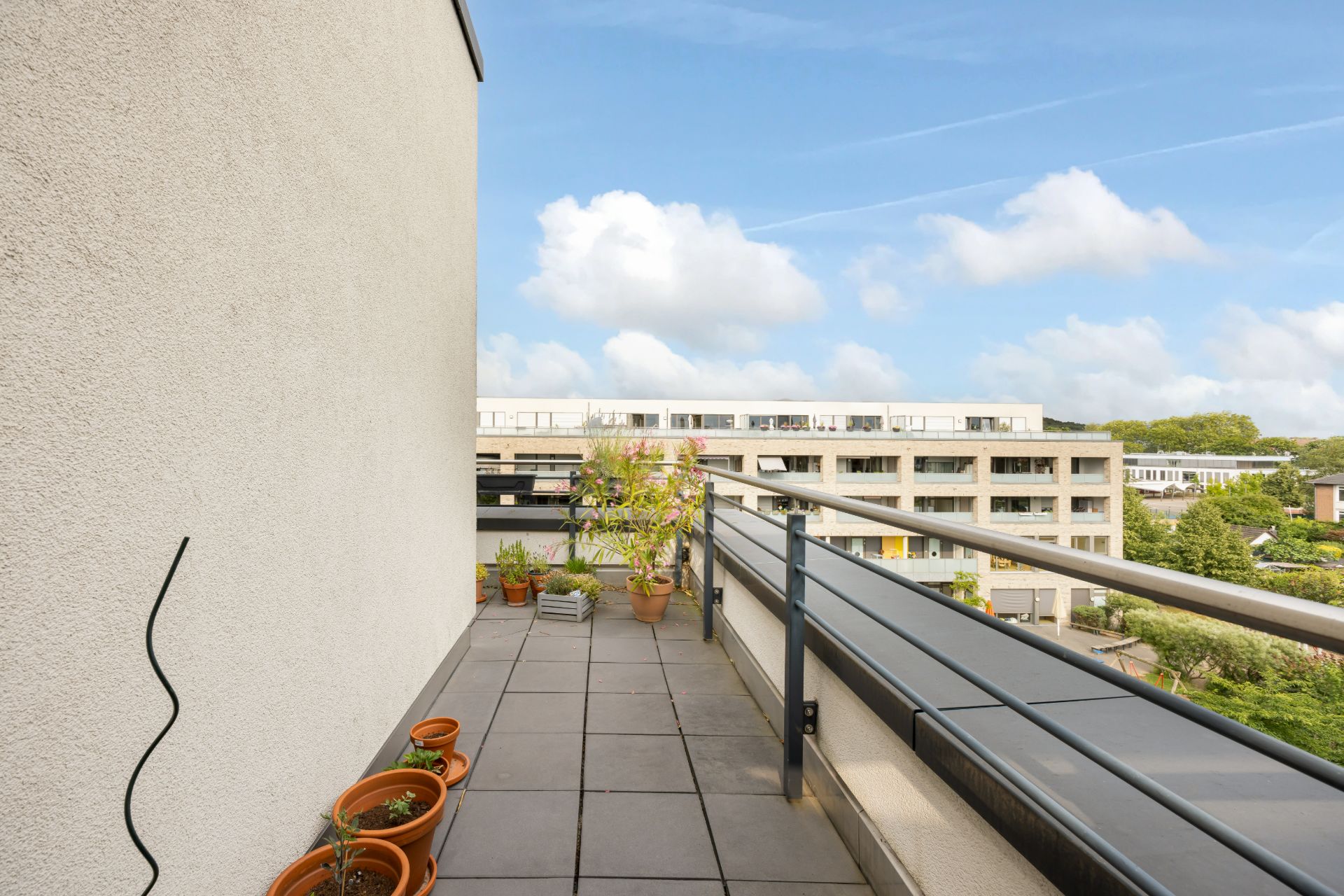 Balkon mit Pflanzentöpfen und Blick auf Wohngebäude, blauer Himmel im Hintergrund.