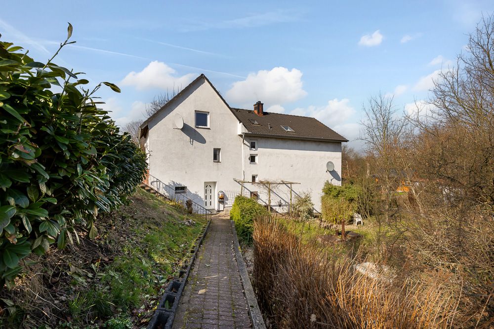 Gepflasterter Weg führt zu einem weißen Wohnhaus mit Satteldach, umgeben von Garten und Büschen.