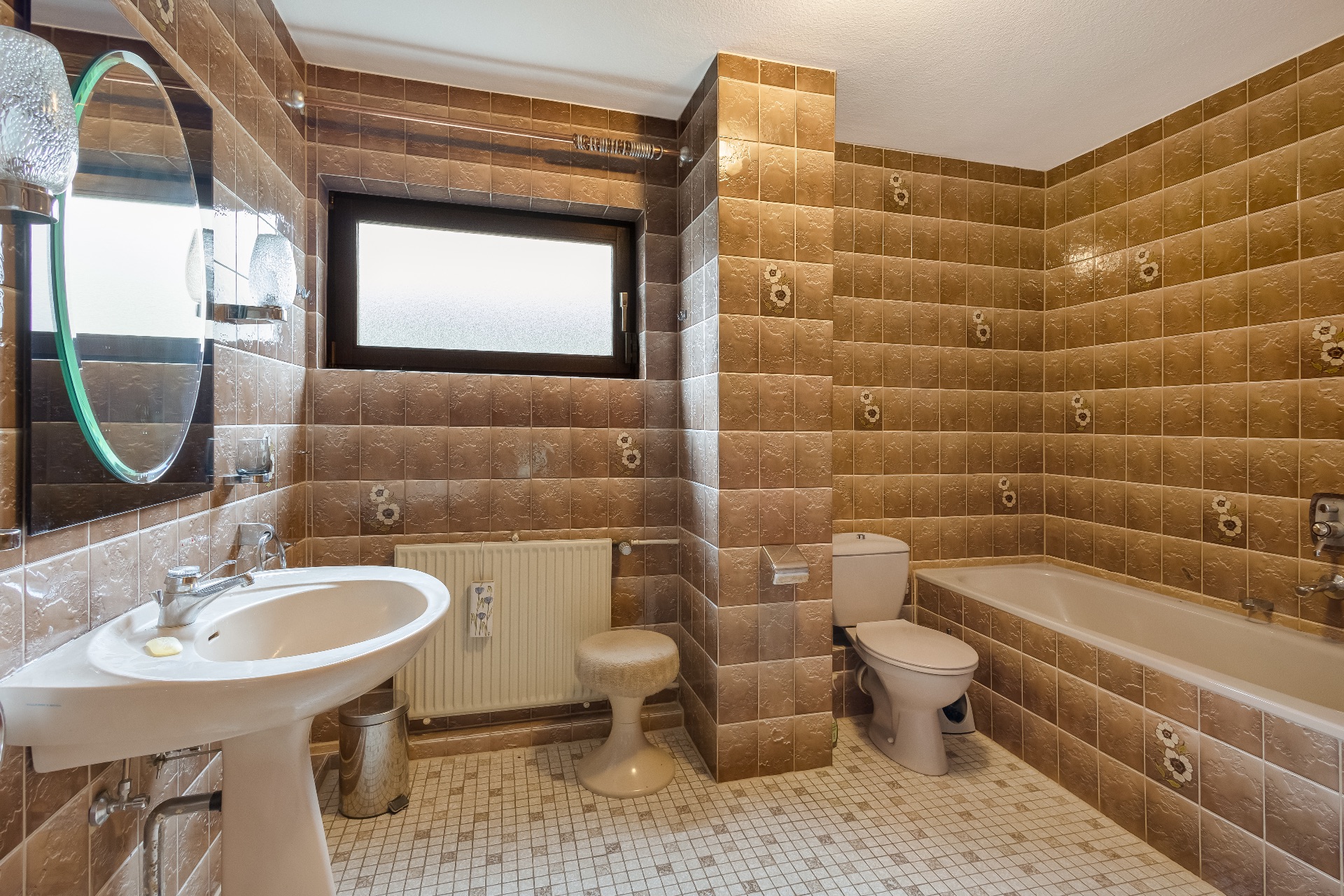 Badezimmer mit braunen Fliesen, Waschbecken, Toilette, Badewanne und Hocker.