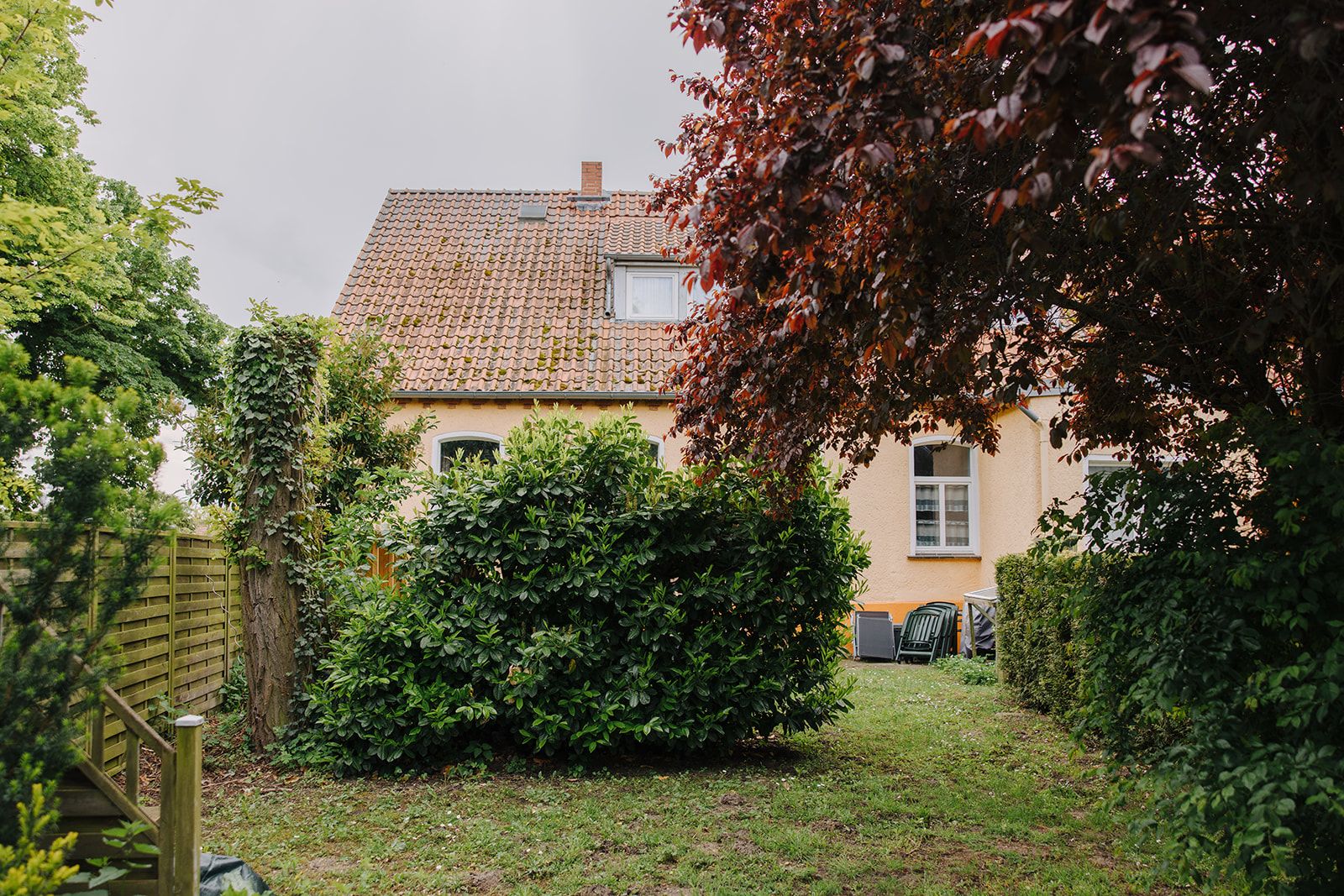 Garten mit Büschen und Bäumen vor einem gelben Wohnhaus mit Dachfenster.
