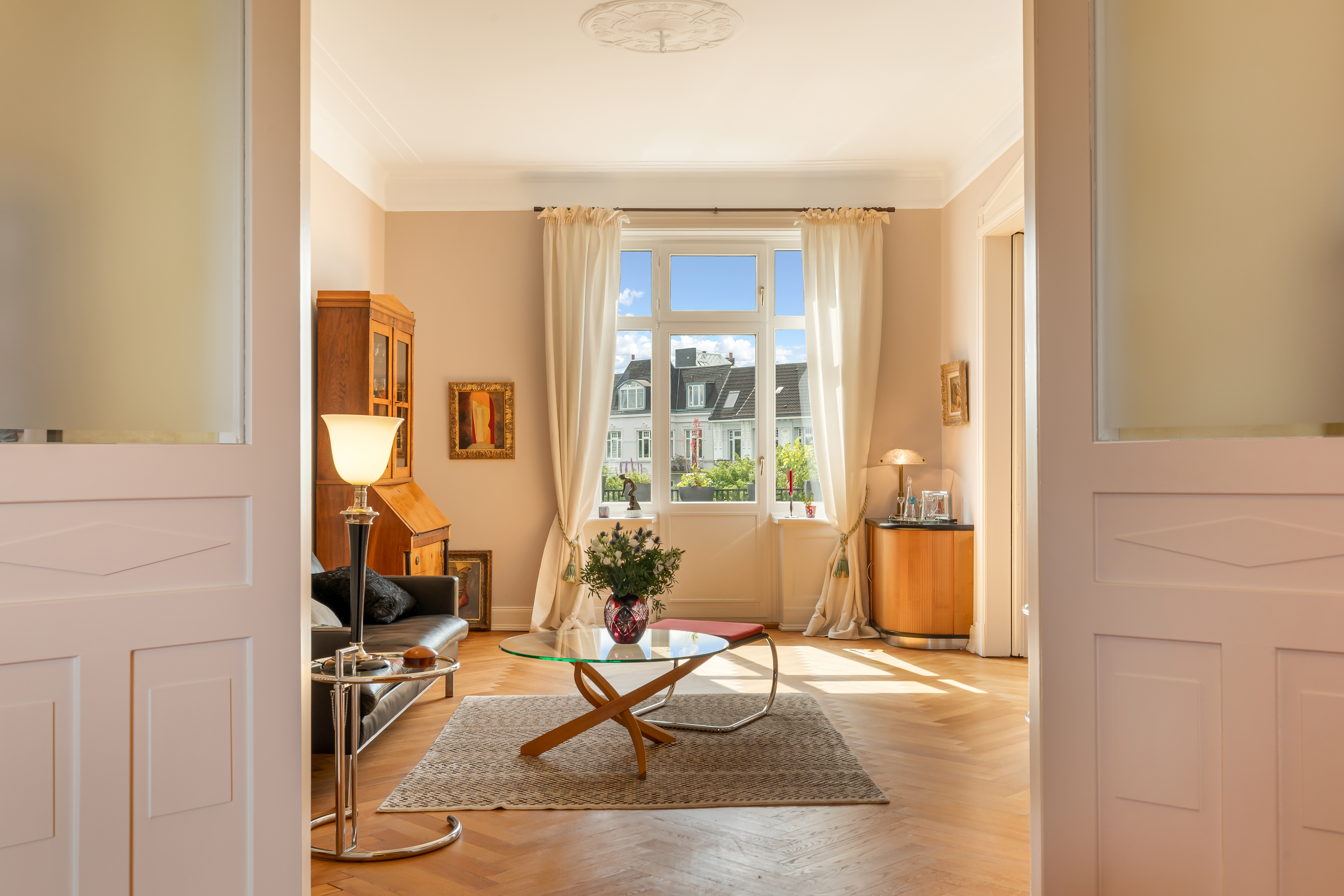 Helles Wohnzimmer mit Sofa, Glastisch, Stehlampe und großem Fenster mit Vorhängen.