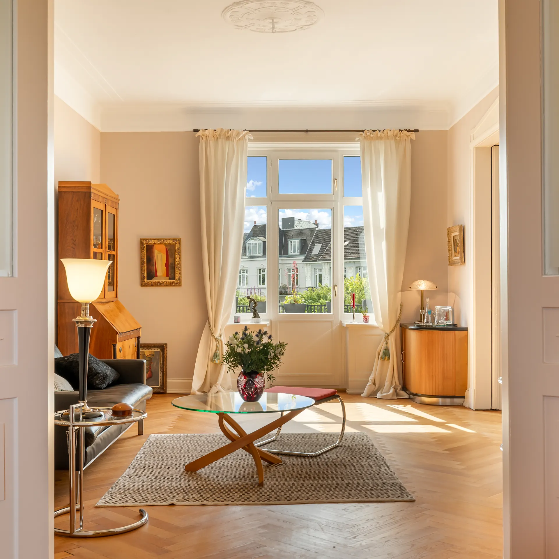 Helles Wohnzimmer mit Sofa, Glastisch, Stehlampe und großem Fenster mit Vorhängen.