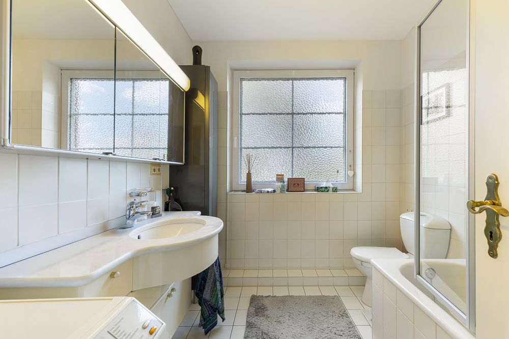 Helles Badezimmer mit Waschbecken, Spiegel, Badewanne, WC und Fenster mit Milchglas.