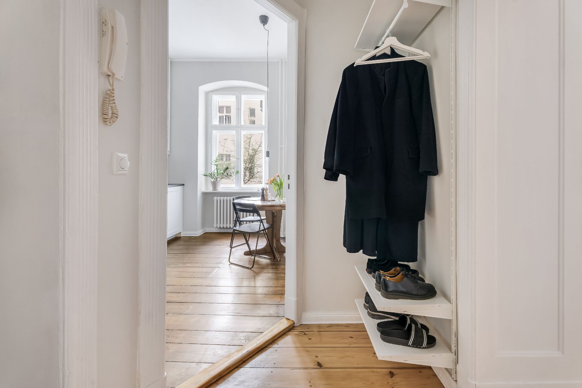Flur mit Garderobe, Schuhen auf Regalen und Blick in ein helles Zimmer mit Tisch und Stuhl.