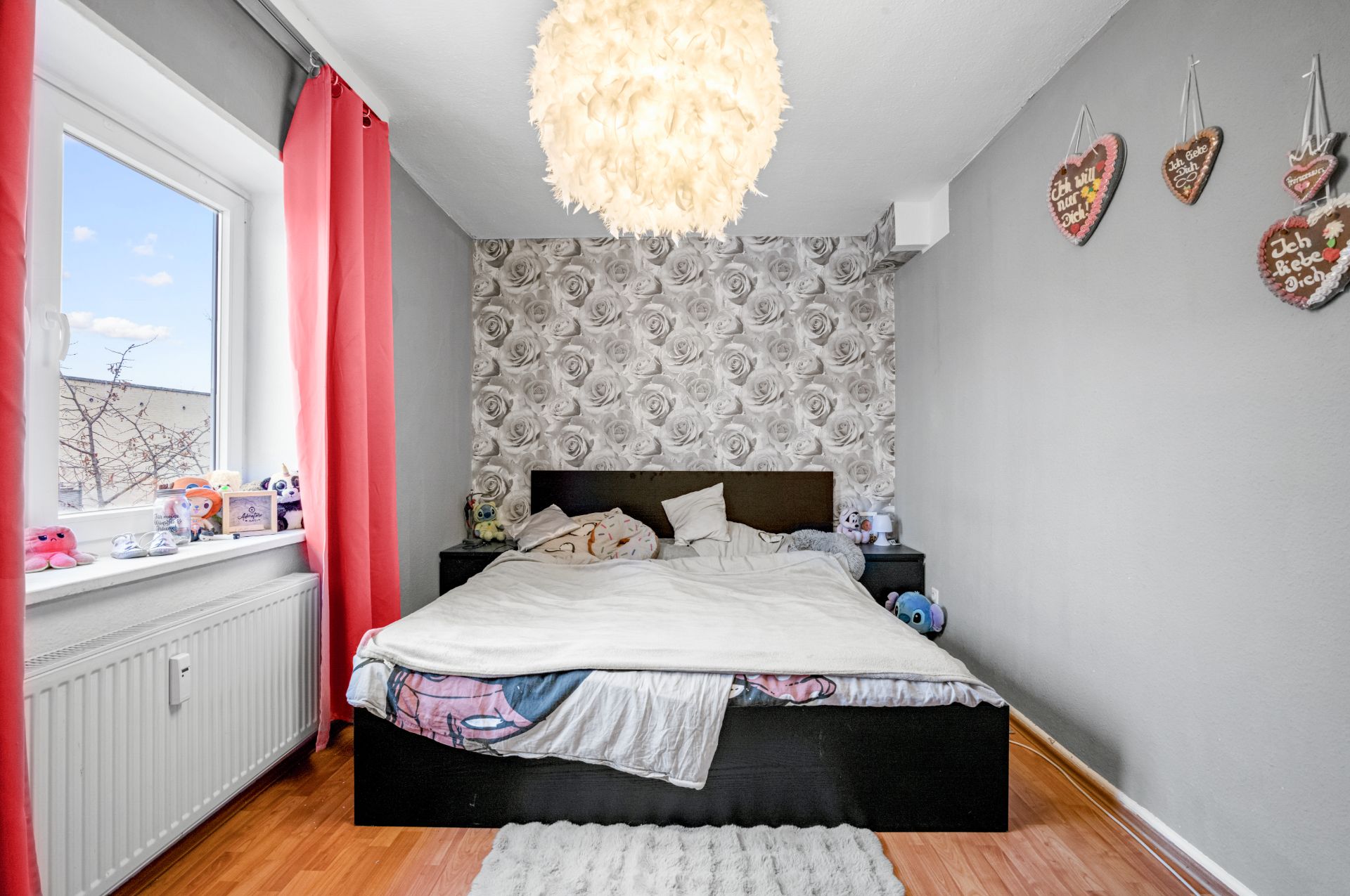 Schlafzimmer mit Doppelbett, rosafarbener Gardine und grauer Rosen-Tapete an der Wand.