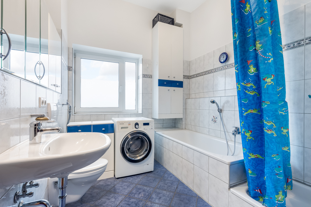 Helles Badezimmer mit Waschbecken, Waschmaschine, Badewanne und blauem Duschvorhang mit Froschmotiven.