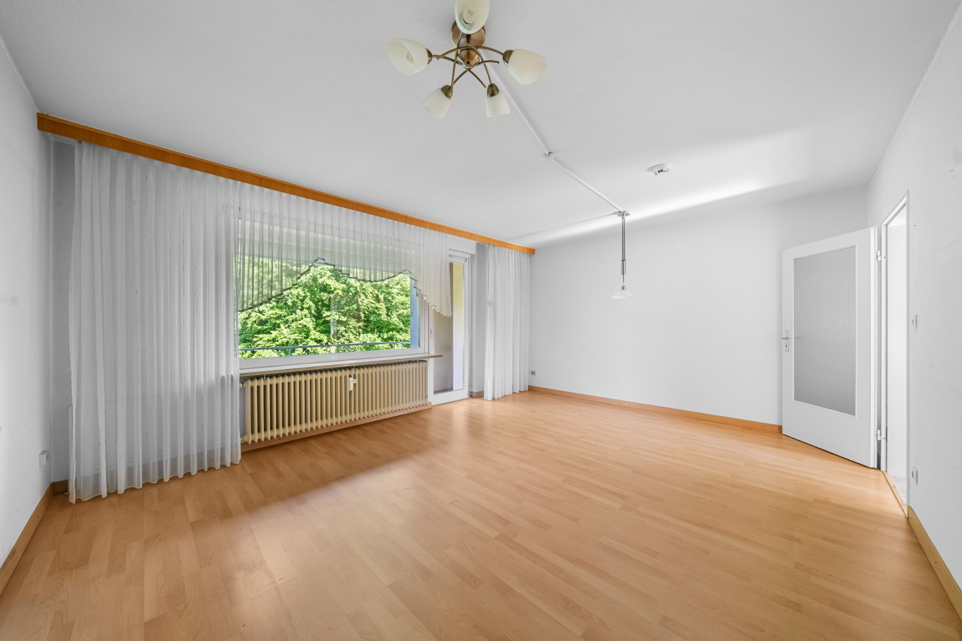 Helles Wohnzimmer mit Holzfußboden, großem Fenster, weißen Wänden und Deckenleuchte.