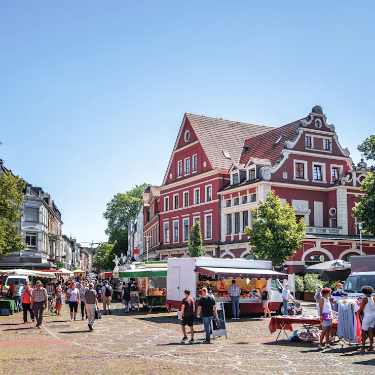 Marktplatz mit Verkaufsständen und einem roten Wohnhaus im Hintergrund bei sonnigem Wetter.