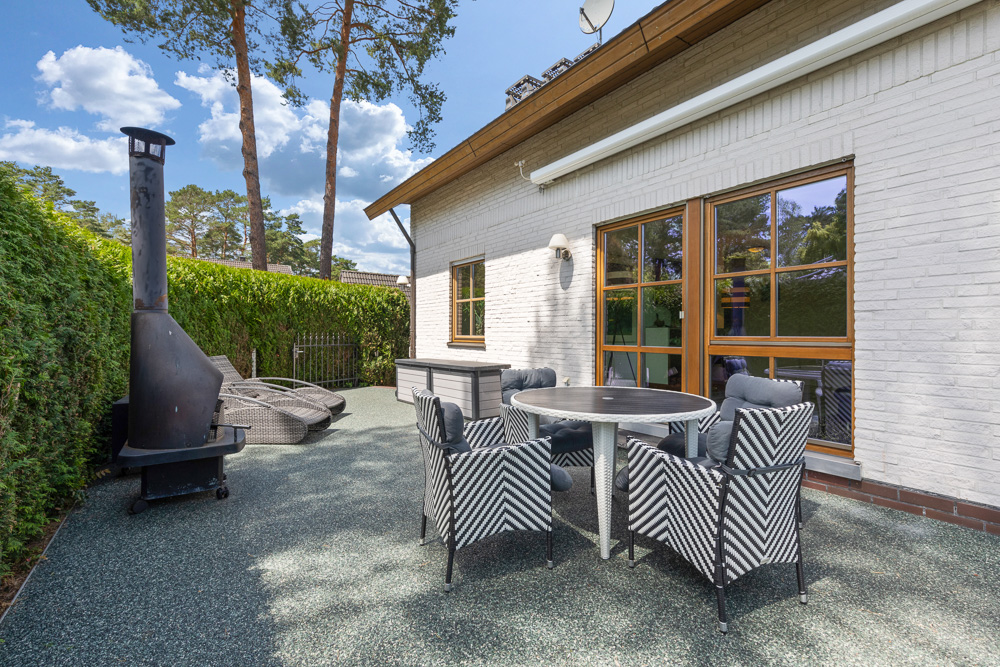Terrasse mit rundem Tisch, vier Stühlen, Kaminofen und Rattansesseln neben einem weißen Haus.
