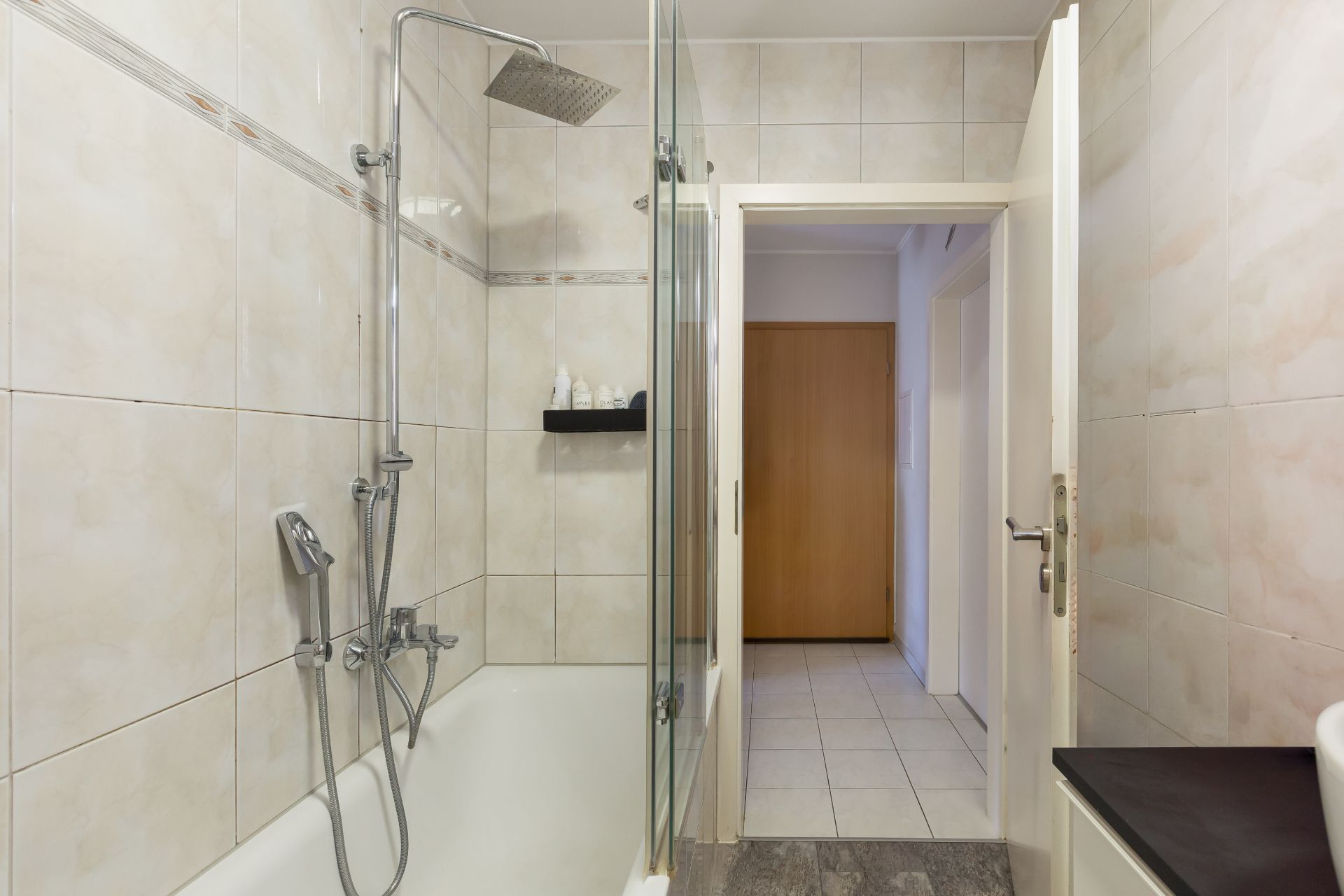 Badezimmer mit Badewanne, Duschkopf, Glasabtrennung und Blick in einen Flur.