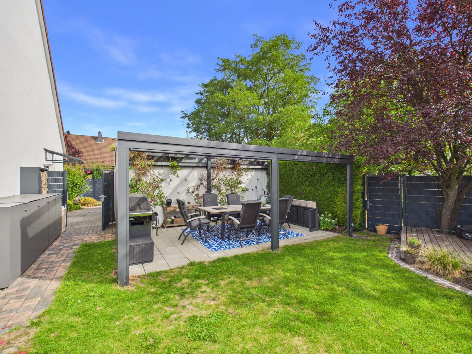 Garten mit überdachter Terrasse, Esstisch und Stühlen, umgeben von Bäumen und Rasen.