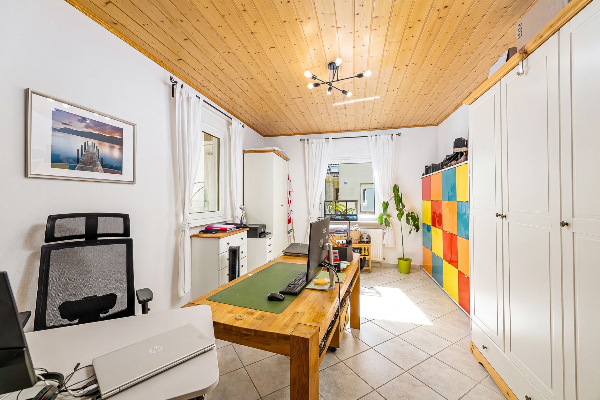 Helles Arbeitszimmer mit Holztisch, Bürostuhl, buntem Regal und Holzdecke.