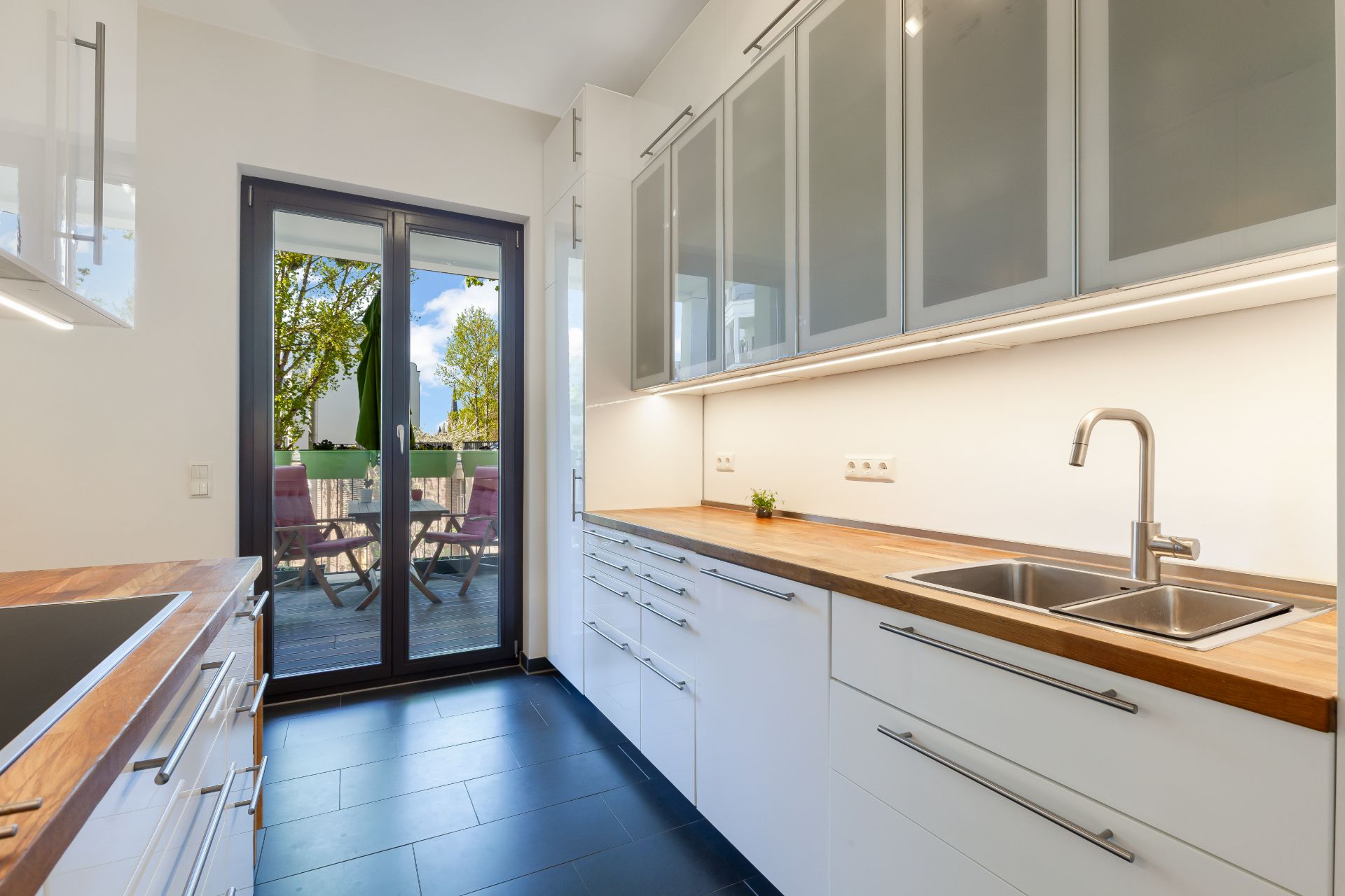 Moderne Küche mit weißen Schränken, Holzarbeitplatten und Zugang zu einer Terrasse.