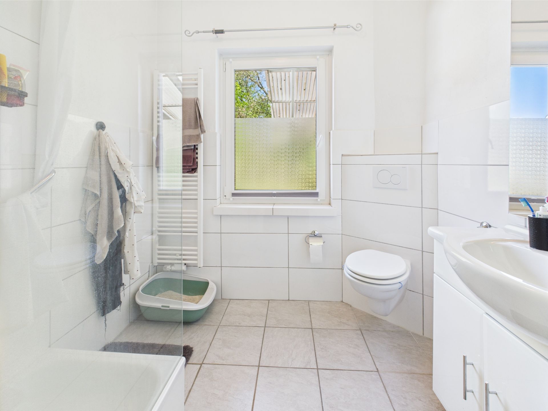 Helles Badezimmer mit wandhängender Toilette, Waschbecken, Handtuchheizung und Katzenklo.
