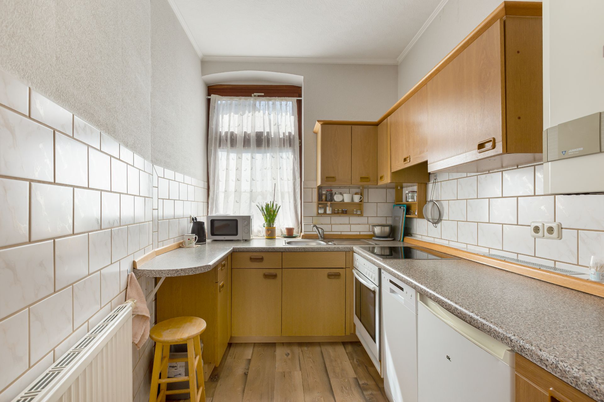 Helle Küche mit Holzmöbeln, weiß gefliesten Wänden, Fenster und Geräten wie Mikrowelle und Herd.