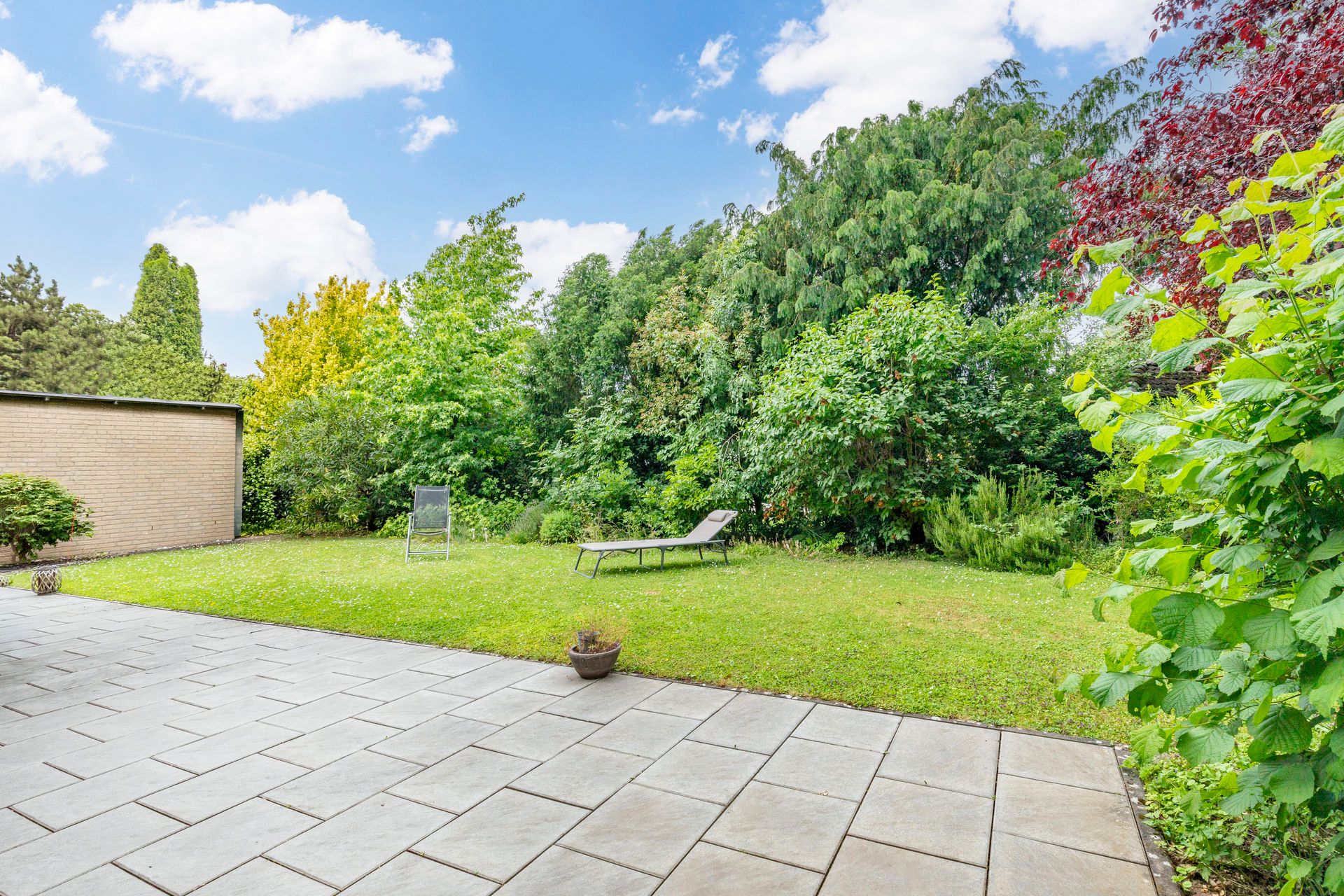 Gepflasterte Terrasse mit zwei Liegestühlen und Rasenfläche, umgeben von dichten Büschen und Bäumen.
