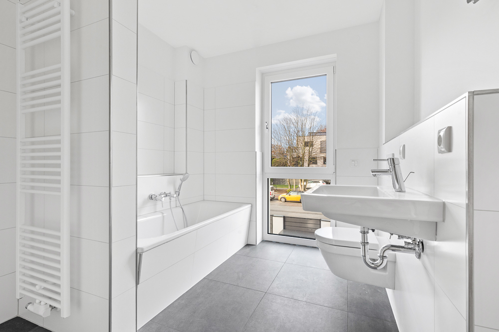 Modernes, weiß gefliestes Badezimmer mit Badewanne, Waschbecken, Toilette und Fenster.